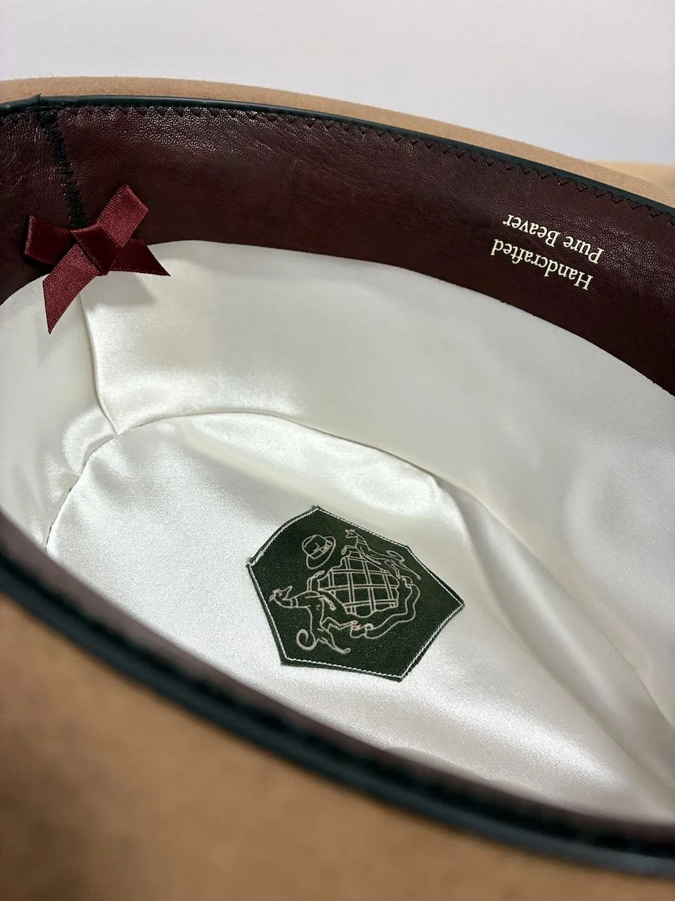 El interior de un sombrero de vestir que muestra un forro de seda blanco con el logotipo de Bolatti Hat Co. en un parche verde oscuro, un lazo granate y letras color marfil que dicen 'Hecho a mano de castor puro' en la badana interior de cuero.