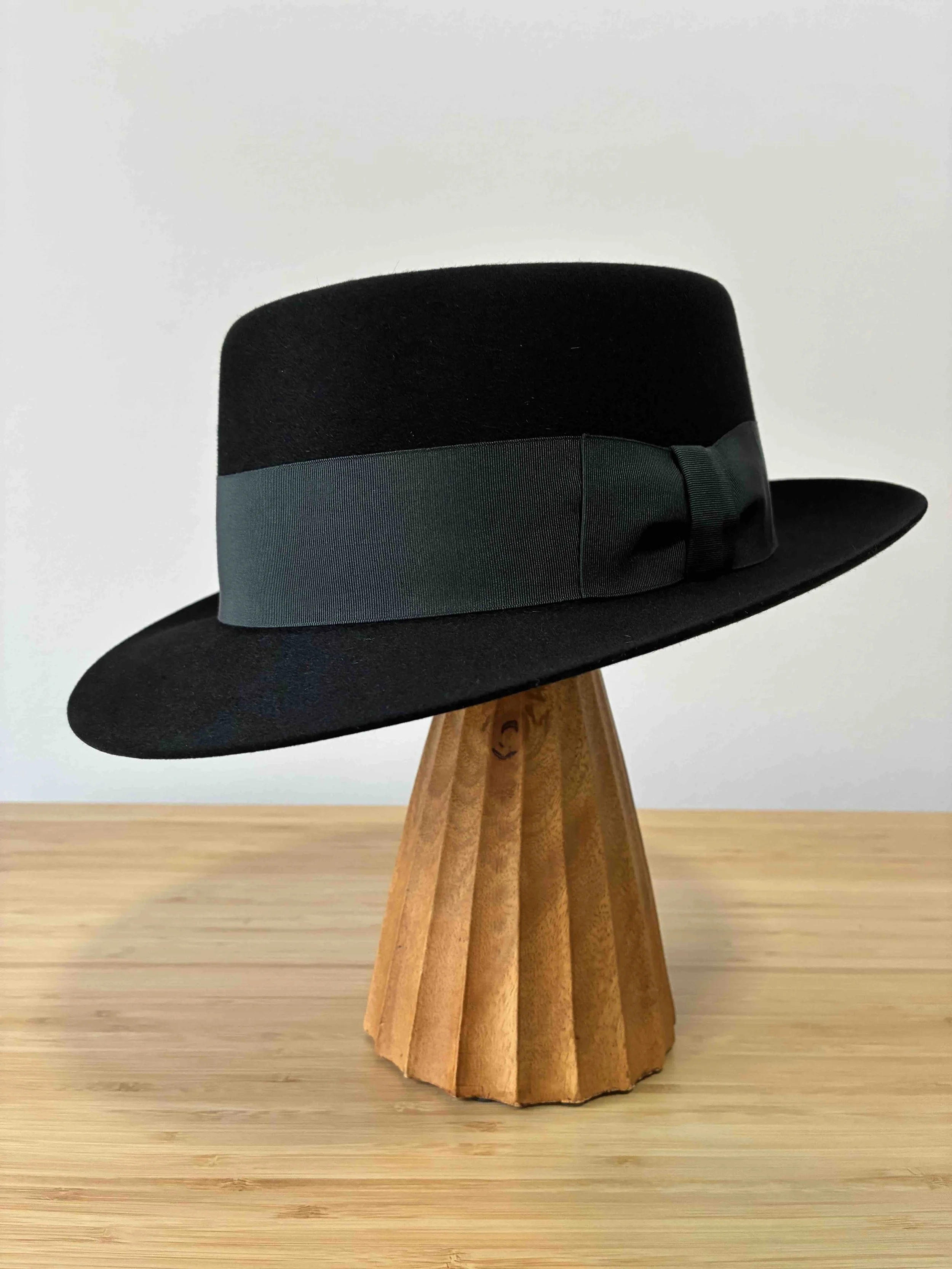 Un sombrero fedora de fieltro negro con una banda ancha de cinta gris y un pequeño lazo decorativo, con ala flexible y copa con forma de pork pie, colocado sobre un soporte de madera, con el ala bajada.