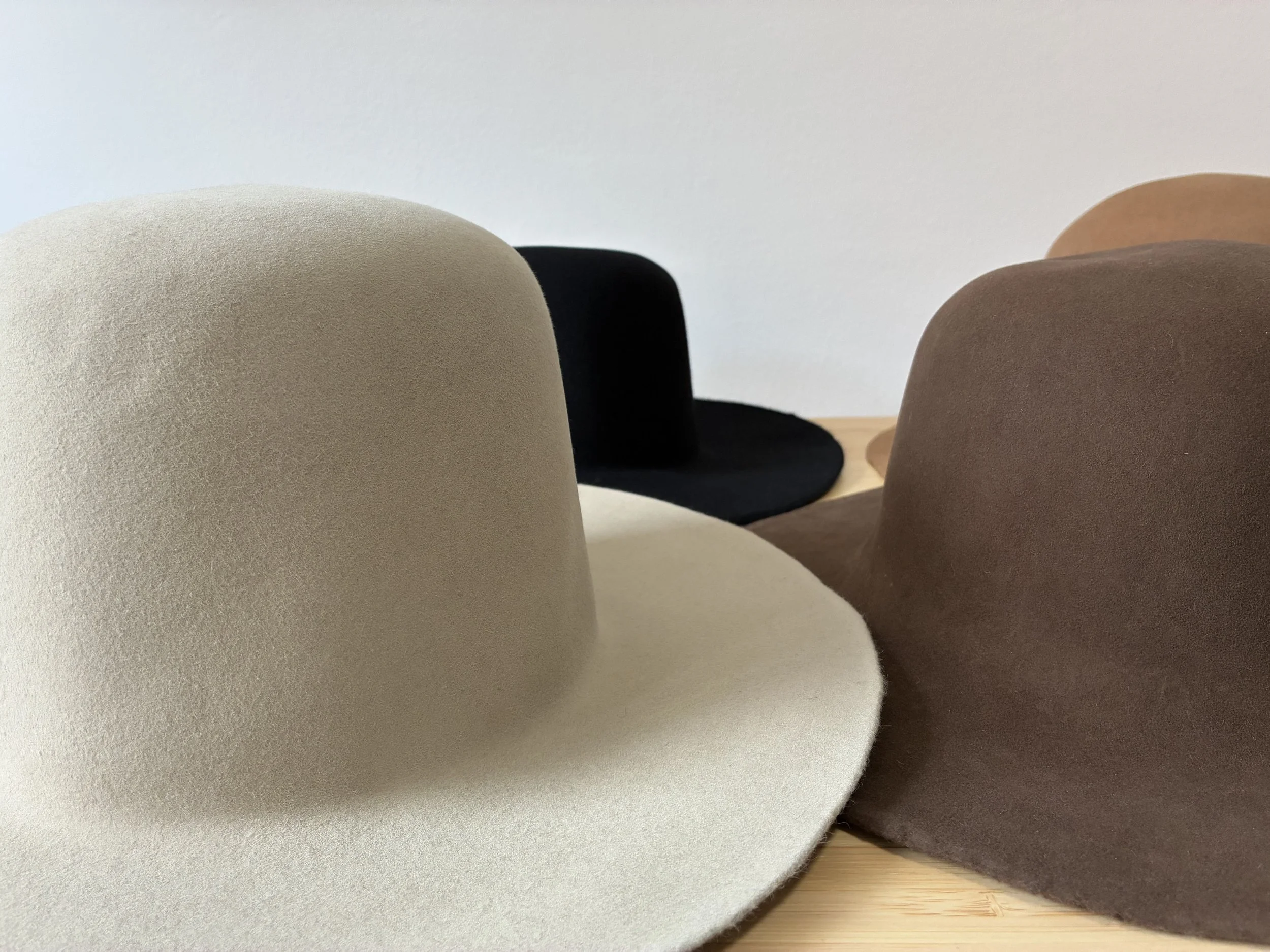 Collection of five felt hat bodies in beige, black, brown, light brown, and dark brown, arranged on a wooden surface against a plain wall.