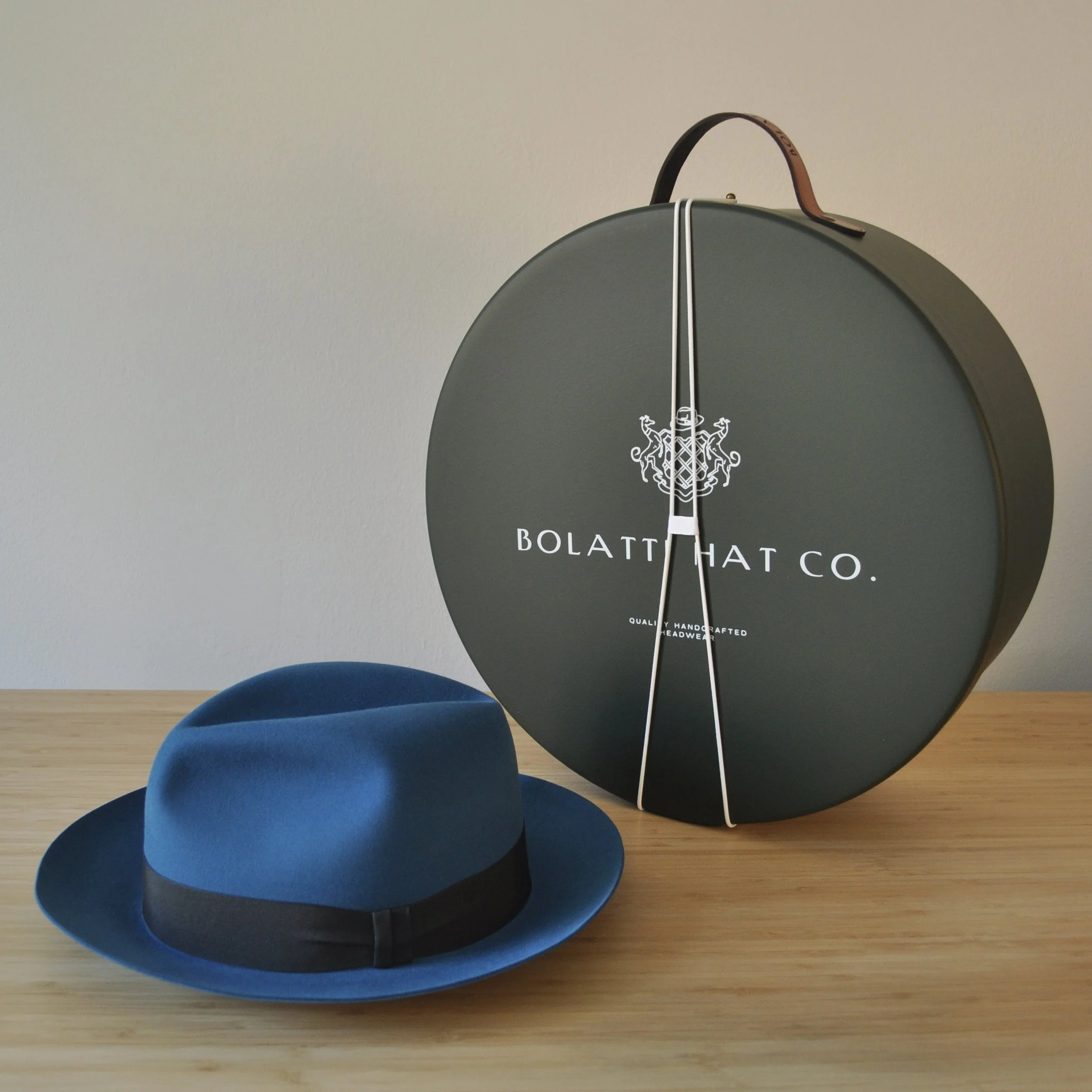 Cobalt blue felt dress hat displayed beside a Bolatti Hat Co. round storage box with leather handle, shown on a wooden tabletop.
