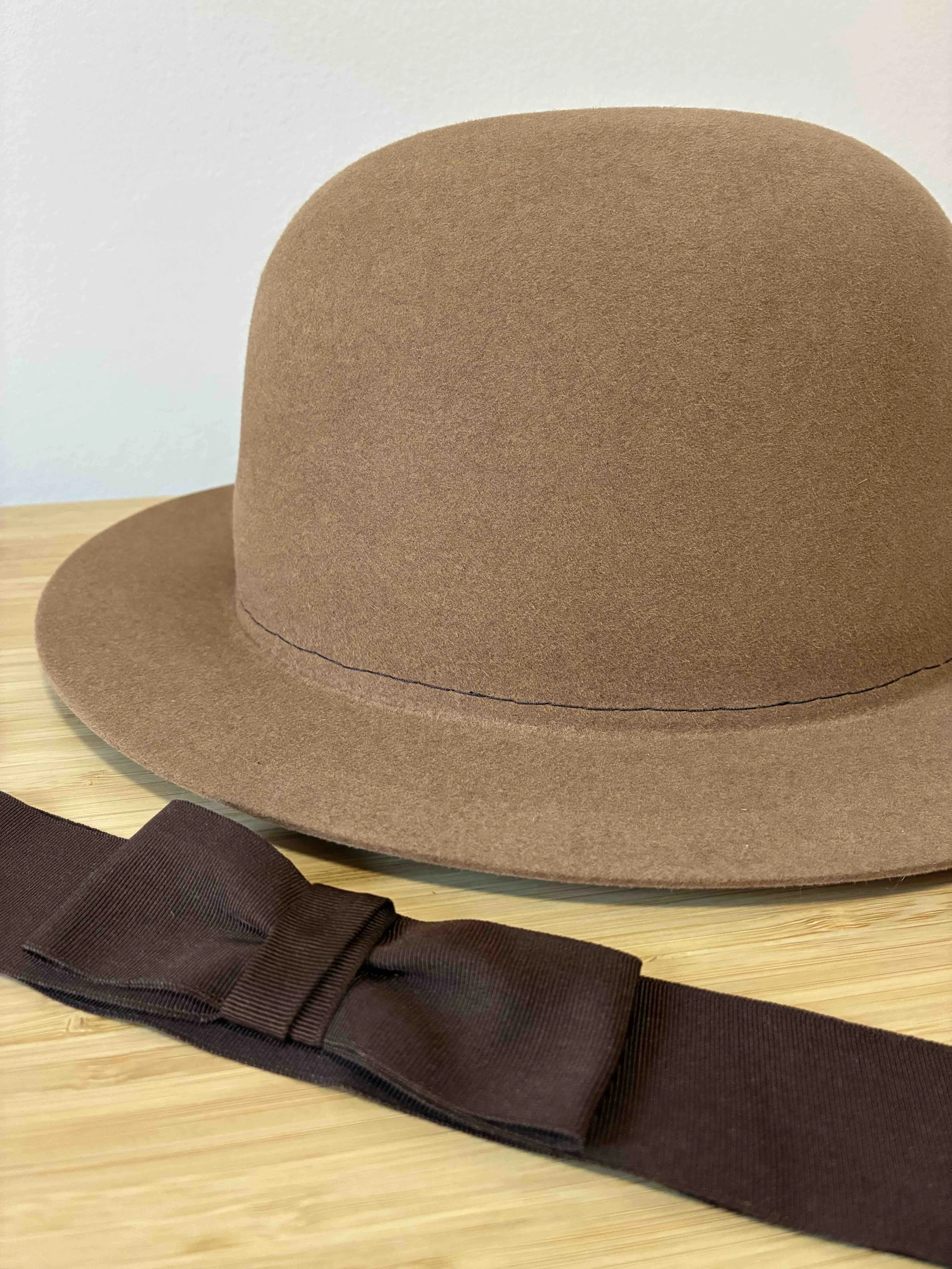 Un sombrero fedora de fieltro de castor color camel sin terminar, sin su banda decorativa de cinta marrón oscuro con un lazo, colocado sobre una superficie de madera clara.