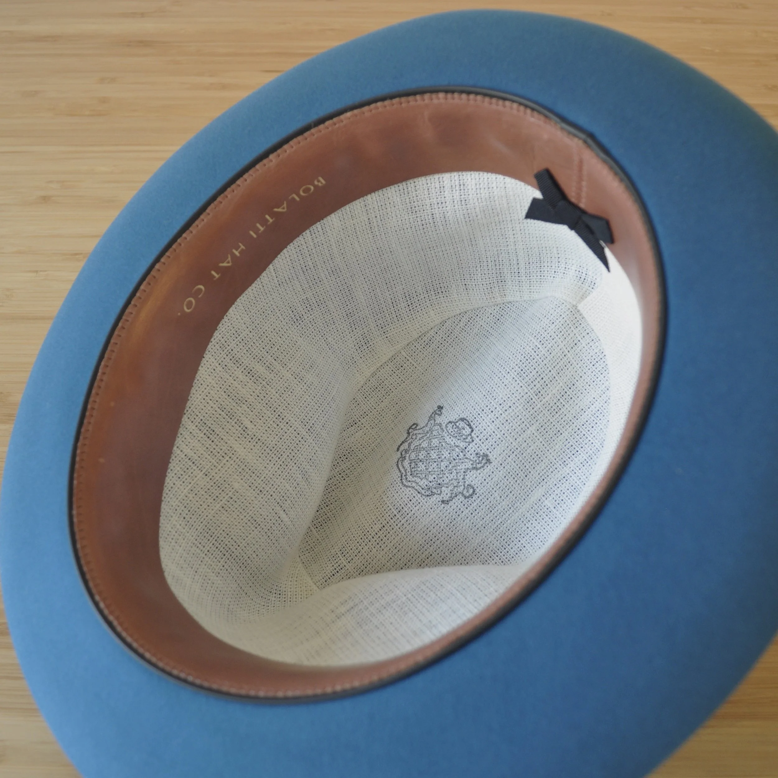 Interior view of a cobalt blue felt dress hat showing a dusty pink leather sweatband, white open weave linen liner, Bolatti Hat Co. crest, and small black grosgrain bow detail.