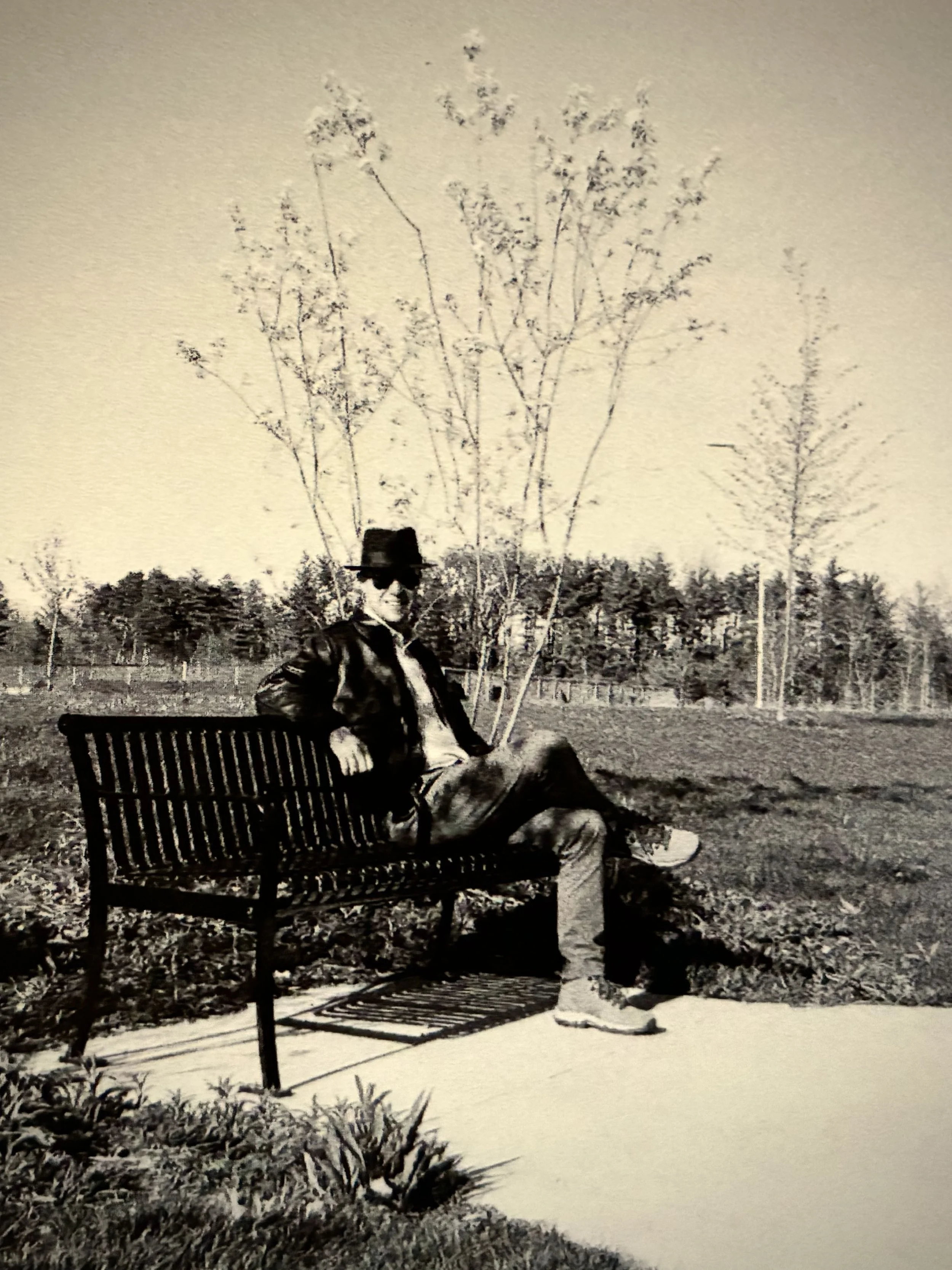 A person wearing sunglasses, a leather jacket, and a hat, sitting on a park bench in a field with a tree in the background.