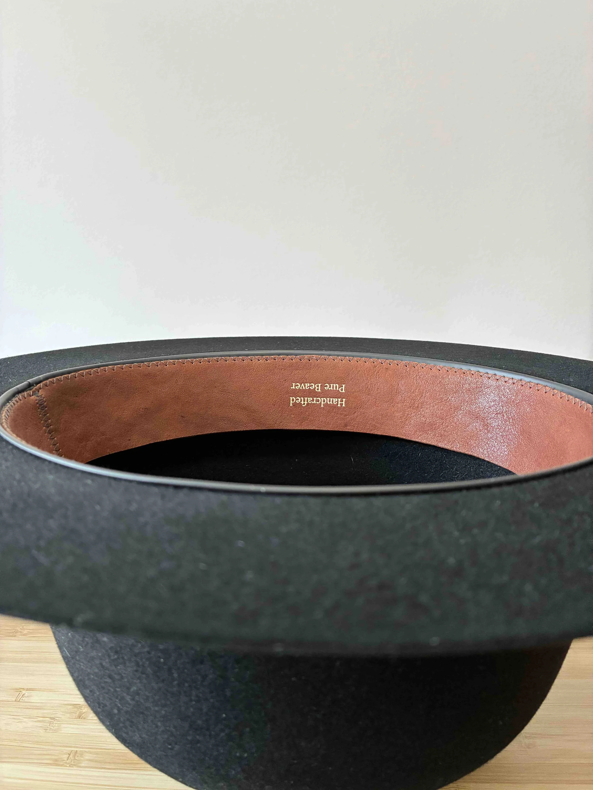 Primer plano de un sombrero de fieltro de castor negro colocado boca abajo sobre una superficie de madera contra una pared blanca, con una badana de cuero marrón que dice 'Handcrafted Pure Beaver'.