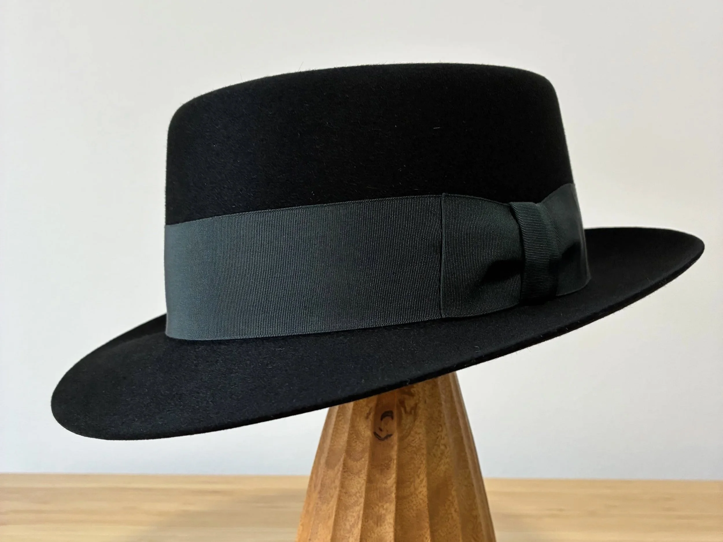 A black felt fedora hat with a wide gray ribbon hat band and a small decorative bow, with snap brim and pork pie shaped crown, placed on a wooden stand, with it's brim snapped down.