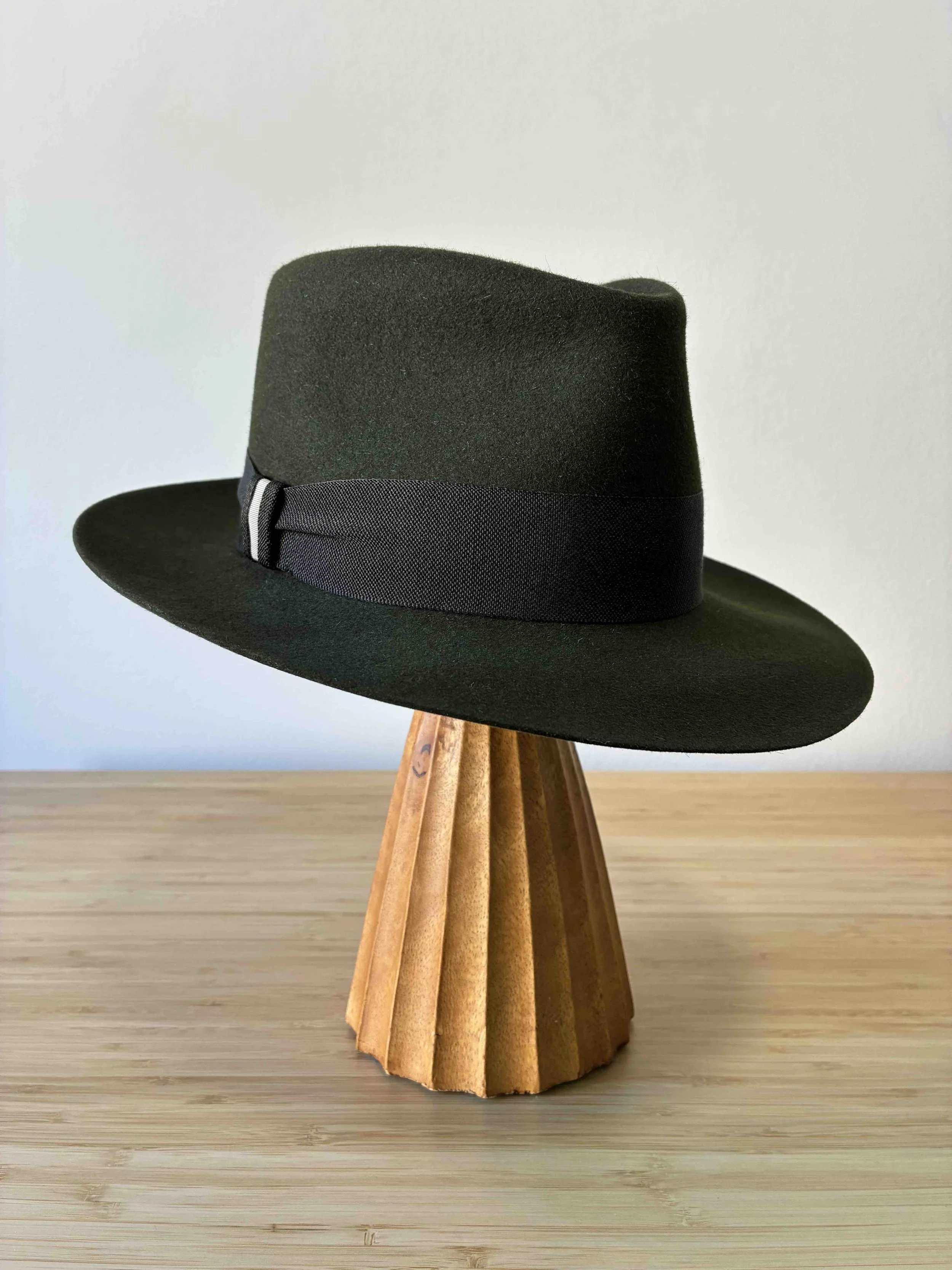 Un sombrero fedora verde oscuro con una cinta de sombrero de textura negra, con ala con caída y una copa en forma de lágrima, colocado sobre un soporte de madera con un fondo liso, y con el ala bajada.