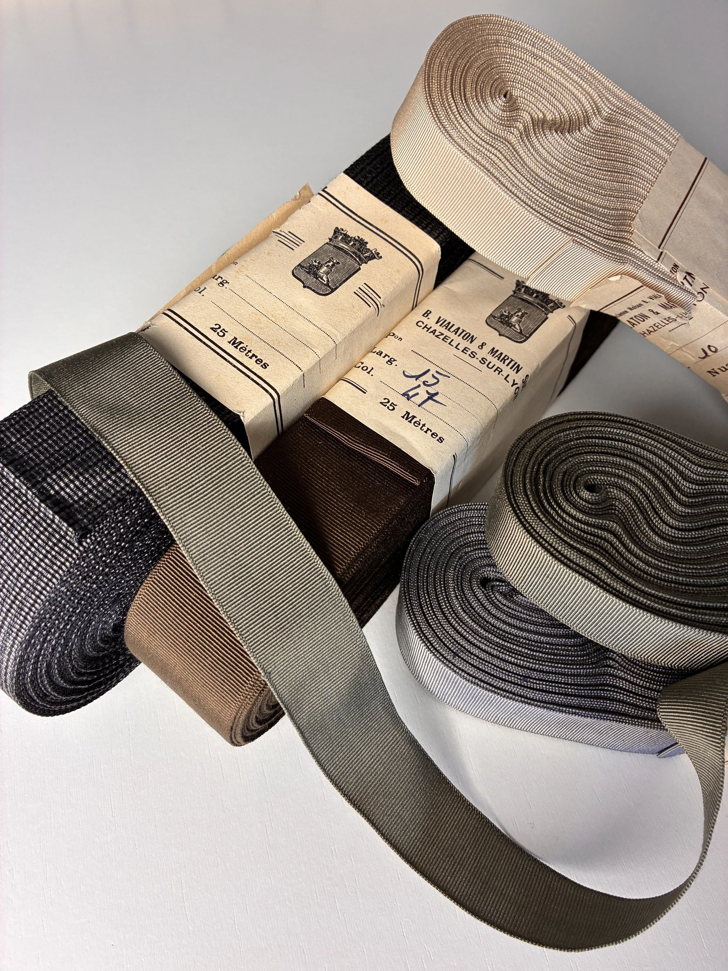 Several rolls of French vintage ribbon in black, beige, silver, gold, and brown, some wrapped in vintage packaging with French text and crest, placed on a white surface.