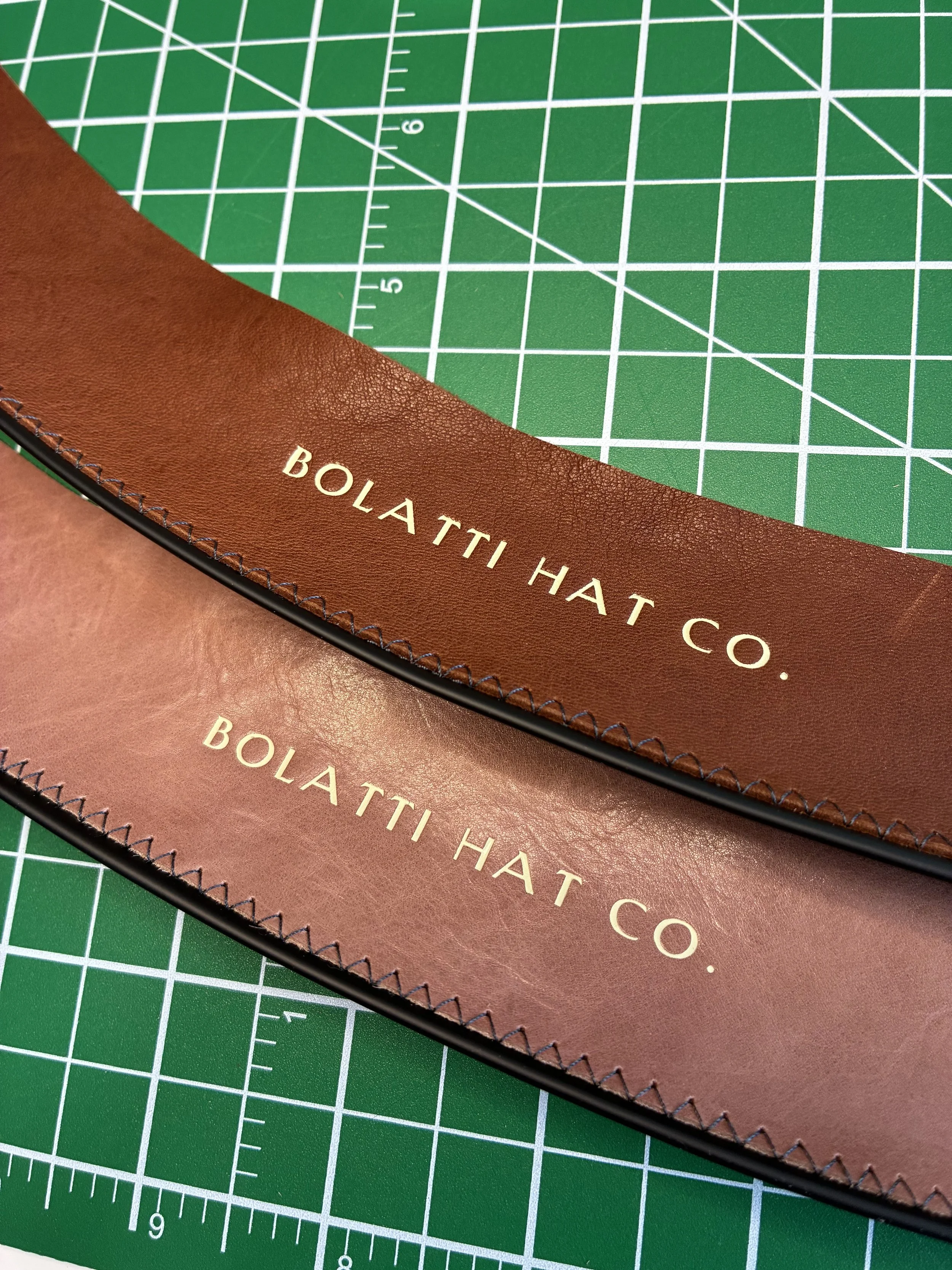 Two leather sweatbands with the text 'BOLATTI HAT CO.' printed on them, placed on a green cutting mat with measurement grid lines.