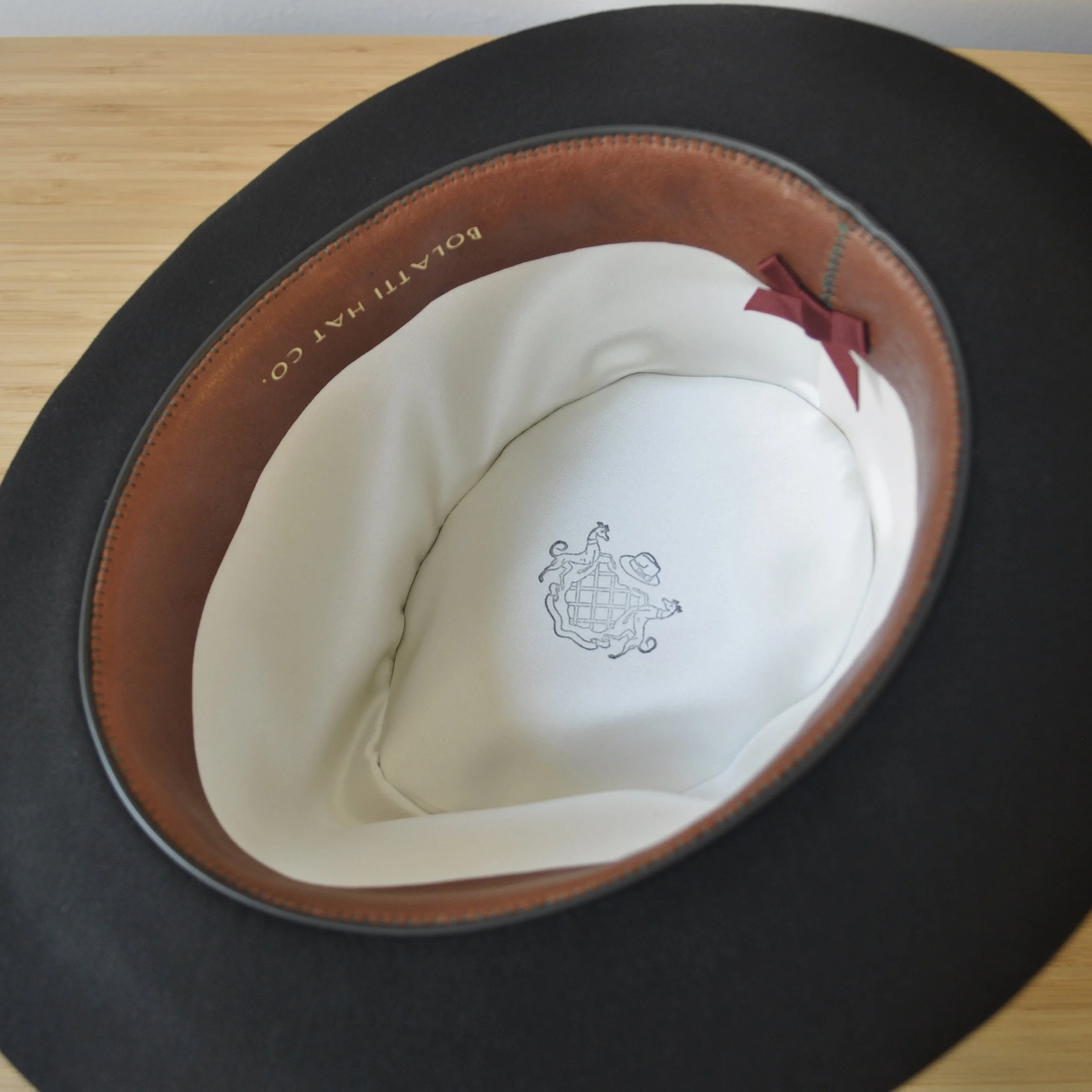Interior view of a black felt dress hat showing a brown leather sweatband, cream liner, Bolatti Hat Co. crest, and small burgundy silk bow detail.