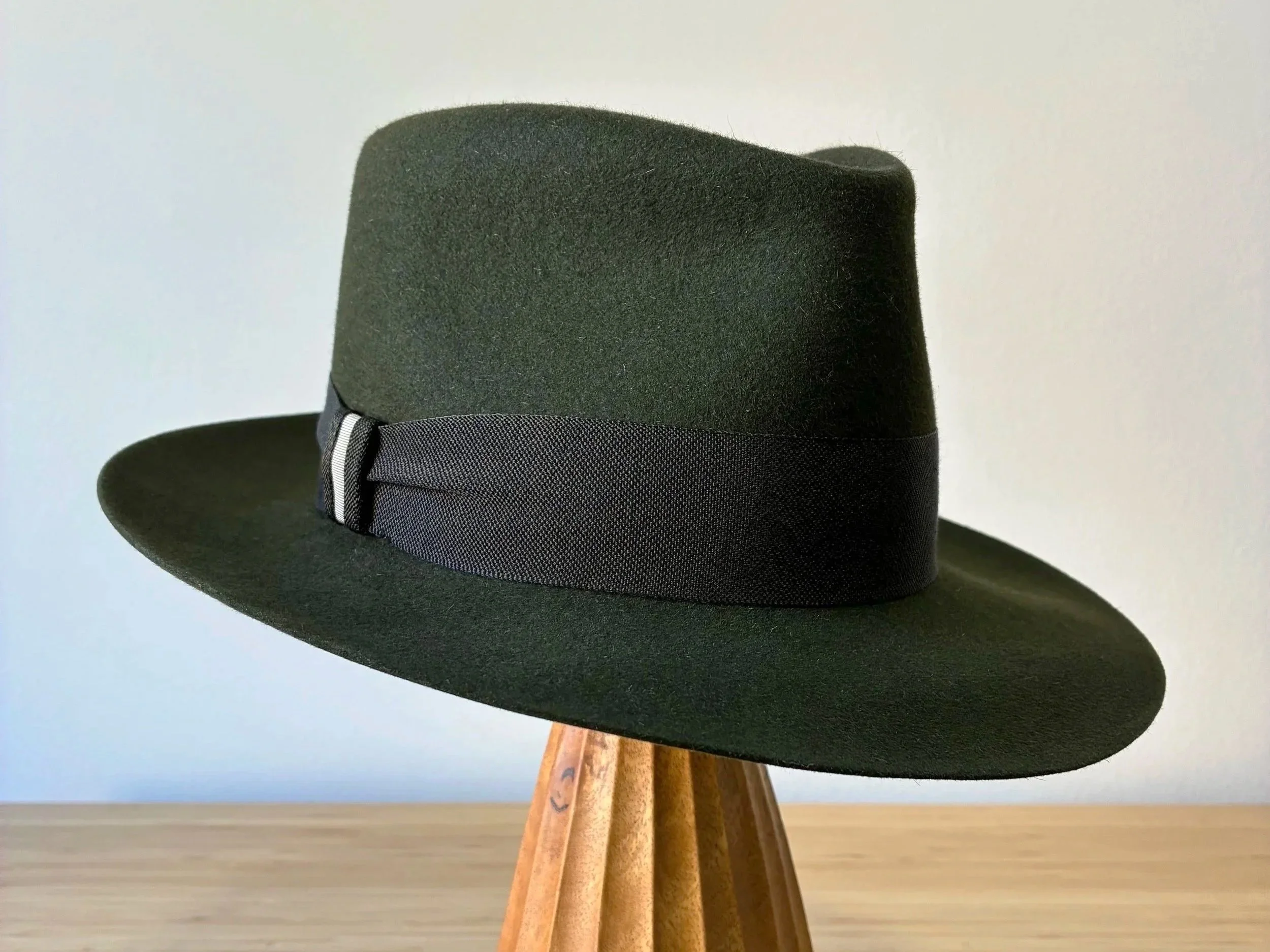 A dark green fedora hat with a black textured ribbon hat band, with snap brim and a teardrop shaped crown, placed on a wooden stand against a plain background, with it's brim snapped down.