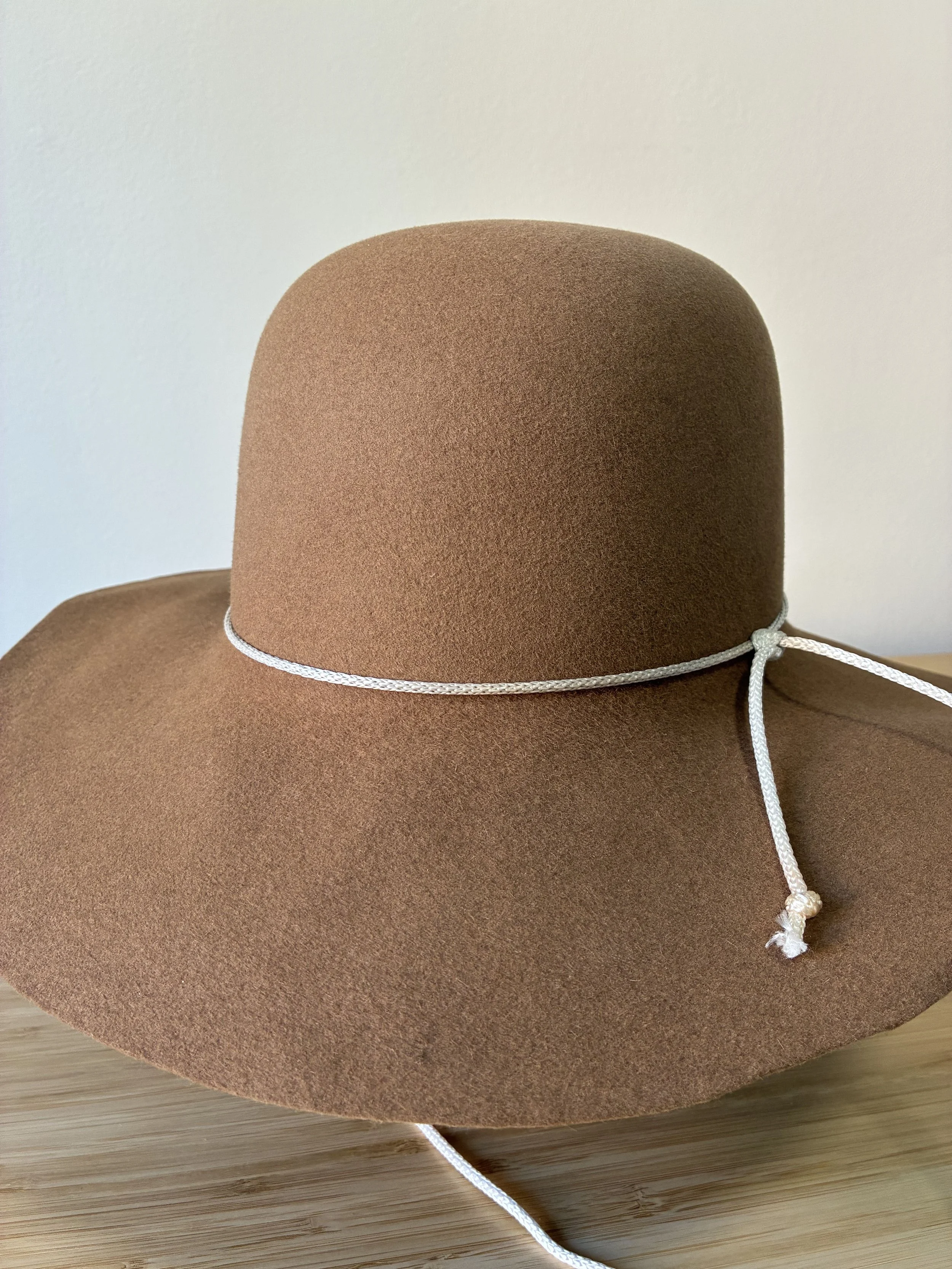 A camel wide-brimmed felt hat body blocked with a white rope around the area where the blocked crown meets the unsnapped brim, sitting on a wooden surface against a white wall.