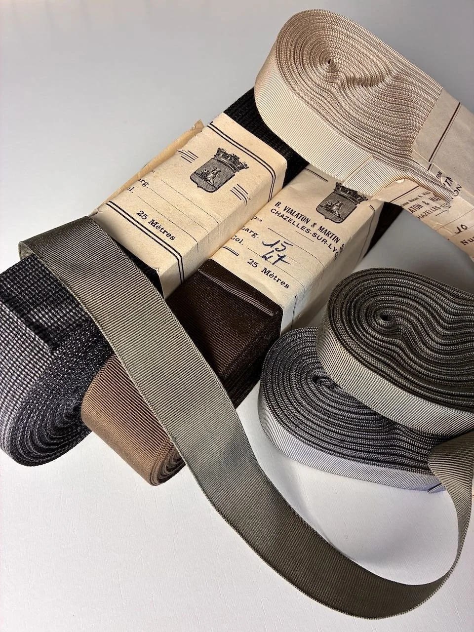 Varios rollos de cinta vintage francesa en negro, beige, plateado, dorado y marrón, algunos envueltos en un embalaje vintage con texto en francés y un escudo, colocados sobre una superficie blanca.