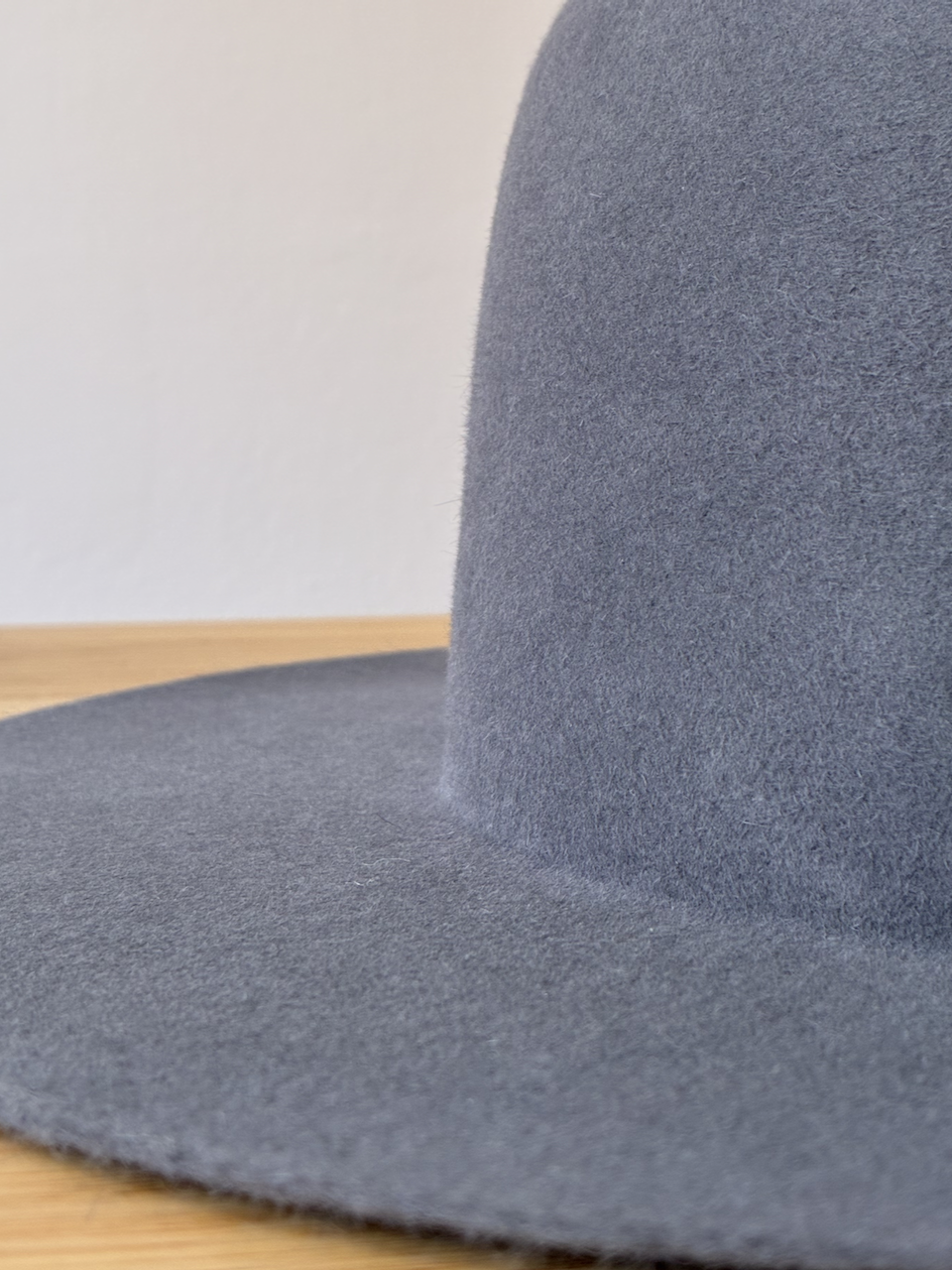 Close-up of a gray felt hat, showing the brim and the crown against a plain white wall.