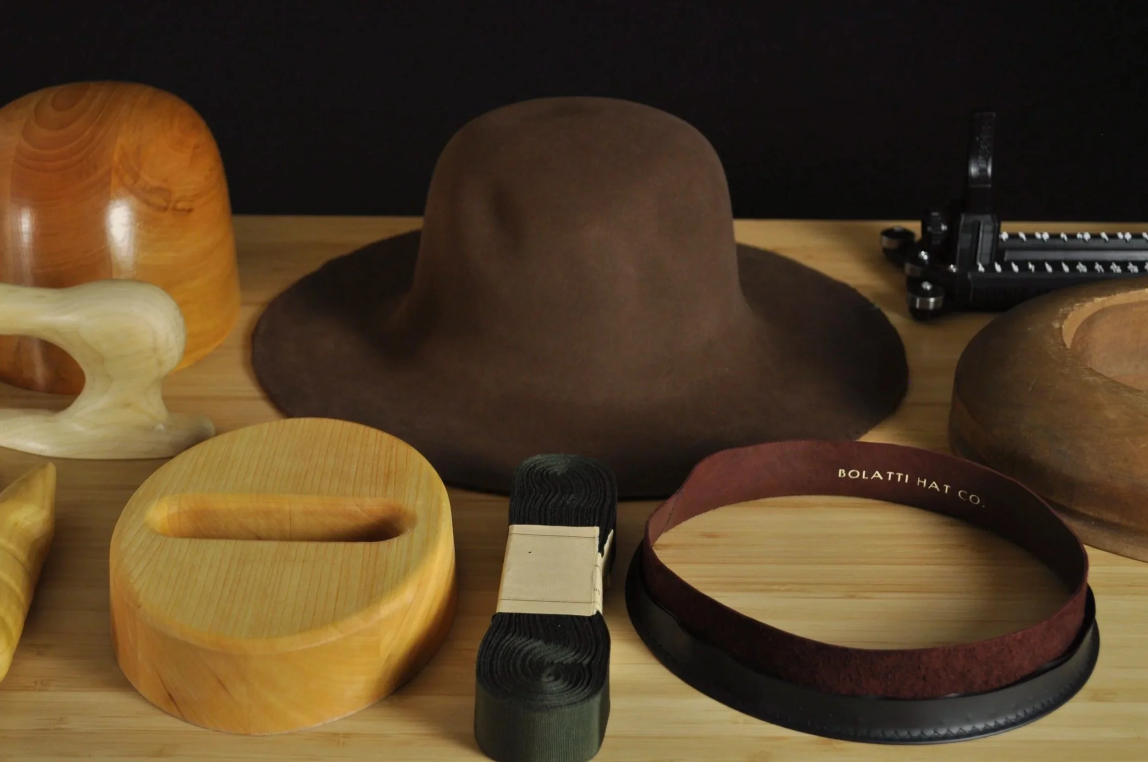 Various wooden and felt accessories including a brown felt hat body, wooden blocks, a maroon leather headband, a dark brown ribbon roll, and a black rounding jack tool on a wooden surface.