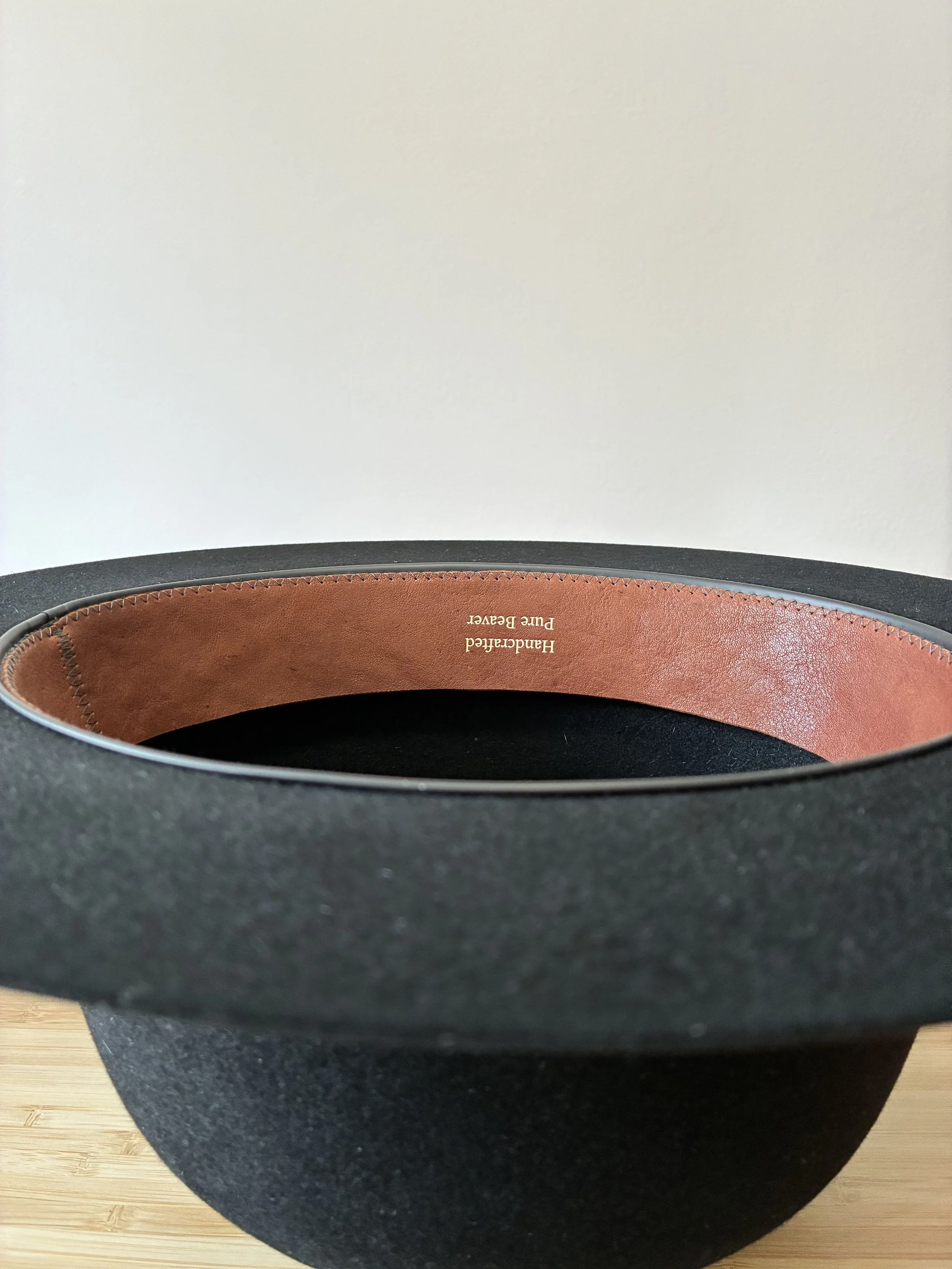 Close-up of a black beaver felt hat sitting upside down on a wooden surface against a white wall, with a brown leather sweatband that reads 'Handcrafted Pure Beaver'.