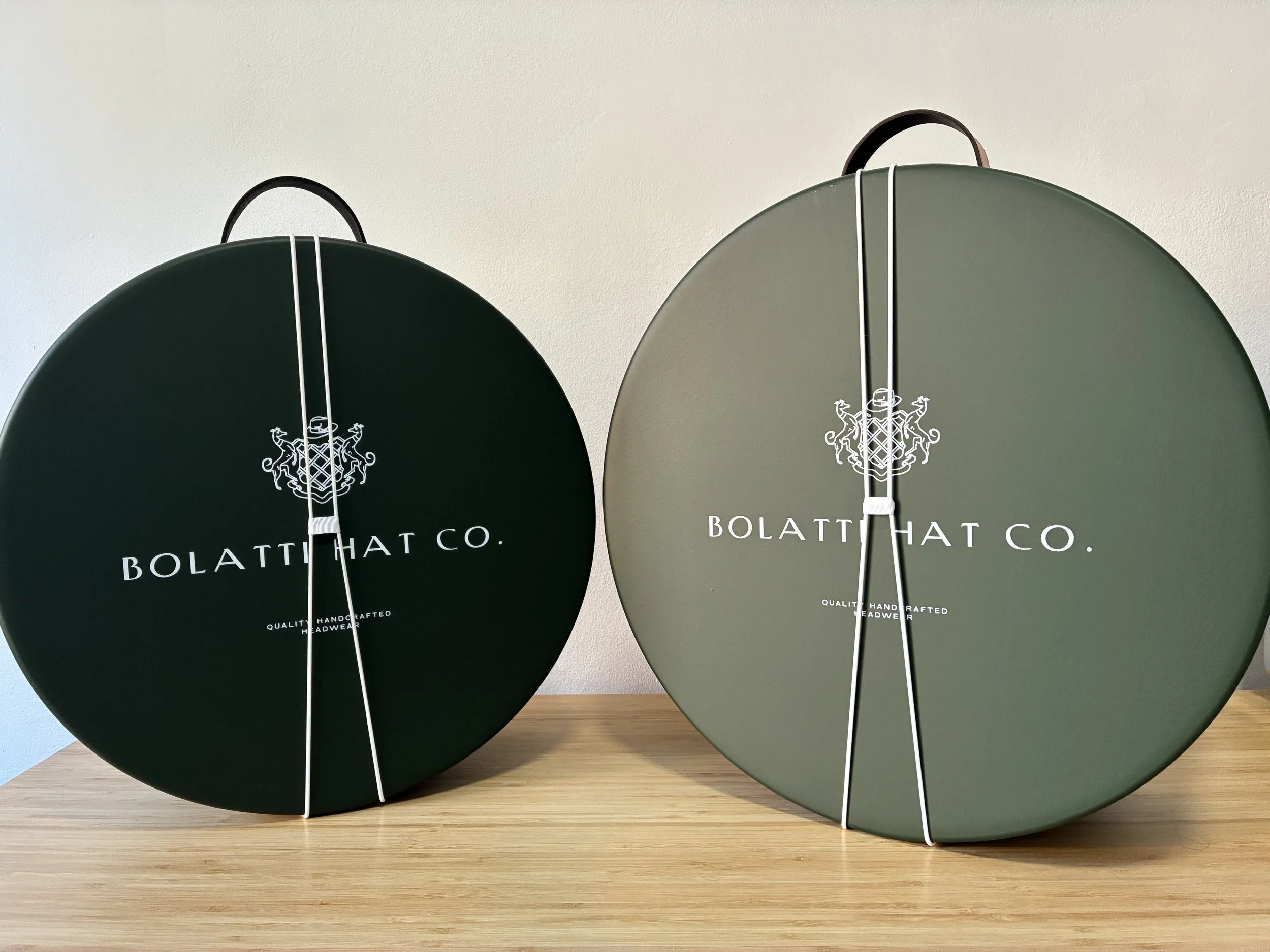 Two round, dark green boxes with white straps, labeled 'BOLATTI HAT CO.' with a crest logo, placed on a wooden surface against a white wall.