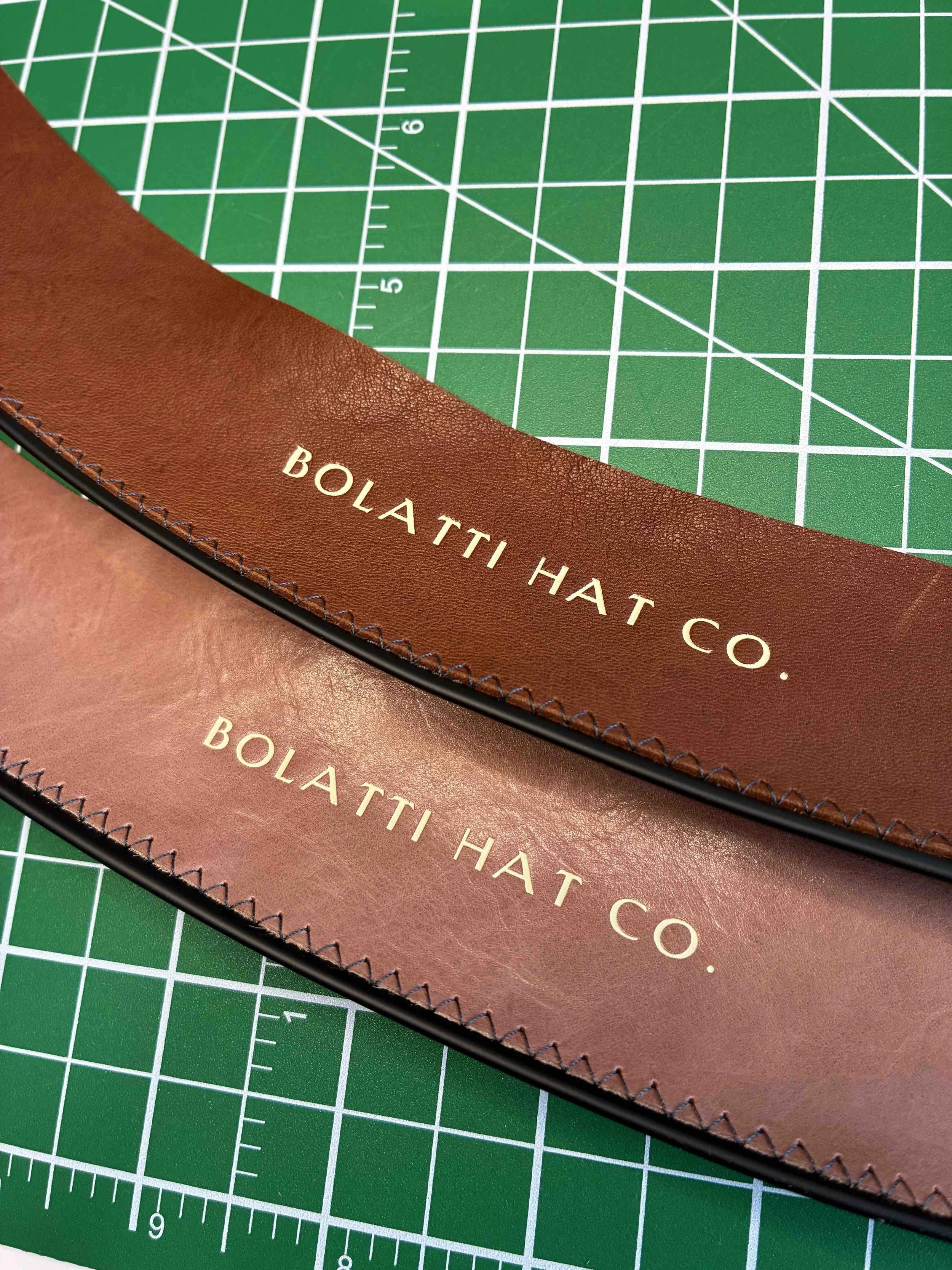 Dos badanas de cuero con el texto 'Bolatti Hat Co.' impreso, colocadas sobre una base de corte verde con líneas de cuadrícula.