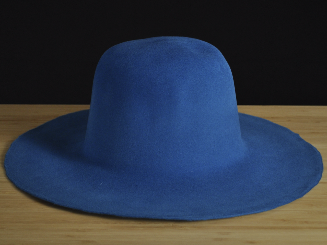 Sombrero de fieltro de pelo de ala ancha de color azul cobalto sobre una superficie de madera, con un fondo negro.