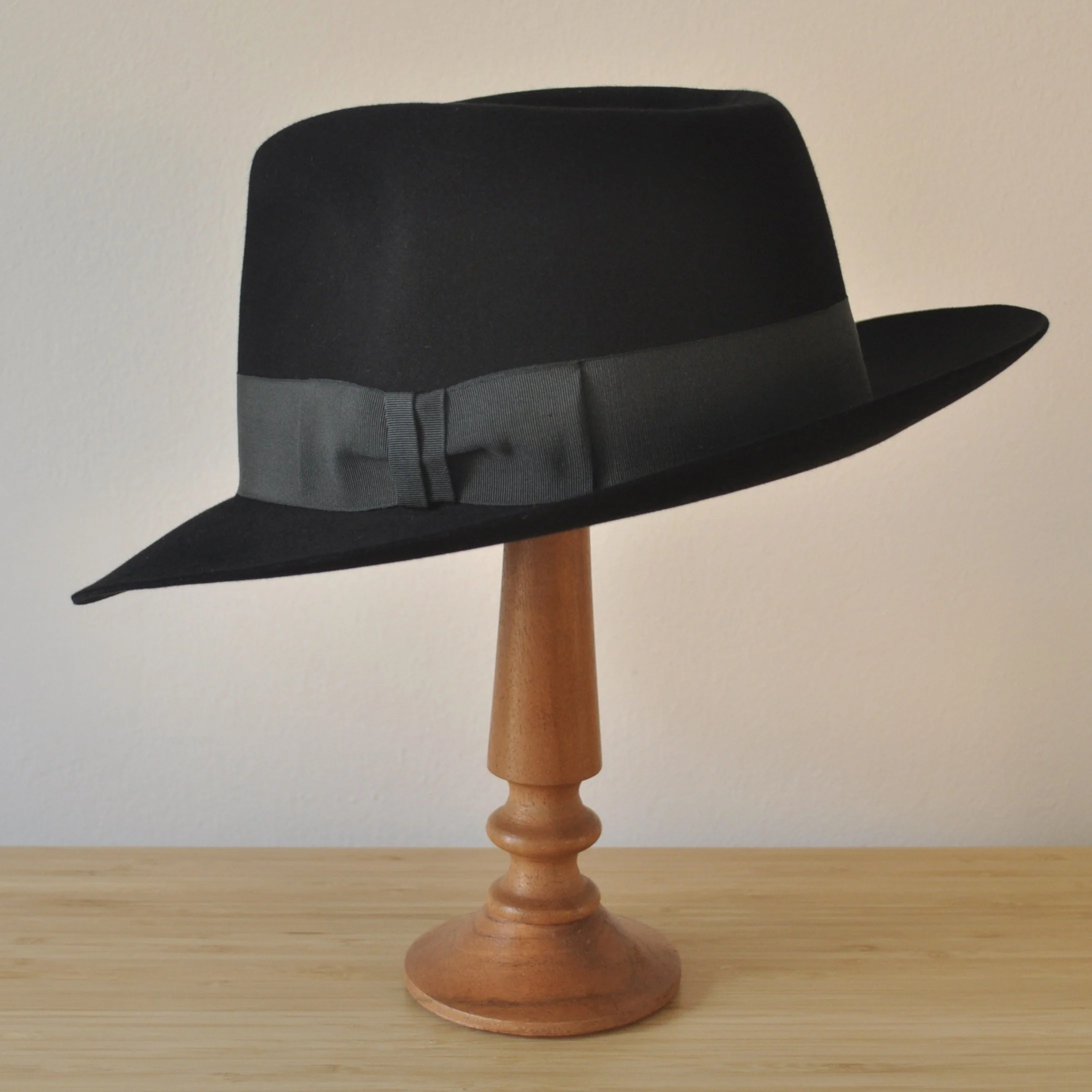 Three-quarter back left view of a black felt fedora with a clean structured crown, medium snap brim, and medium-dark bluish green grosgrain ribbon with classic bow.