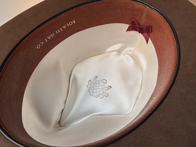 The interior of a dress hat showing an ivory coloured silk lining with Bolatti Hat Co. crest, a maroon bow, and ivory lettering that says ‘Bolatti Hat Co.’ on the inner leather sweatband.