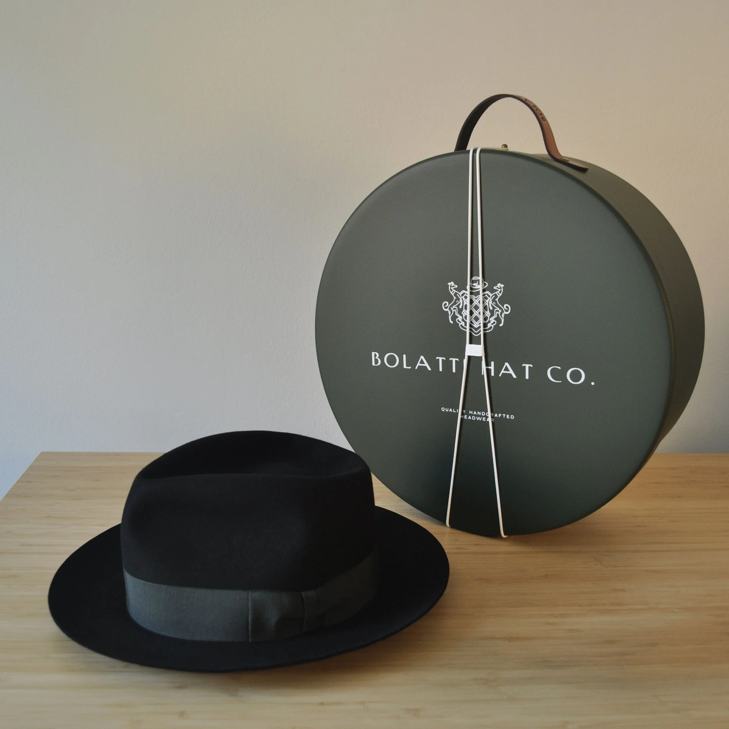 Black felt dress hat displayed beside a Bolatti Hat Co. round storage box with leather handle, shown on a wooden tabletop.