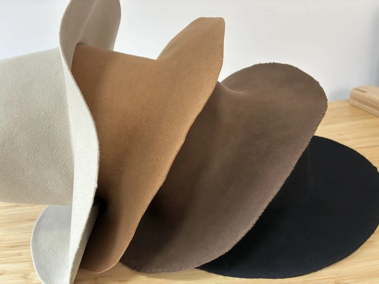 Cuerpos de sombreros de fieltro apilados en colores crema, marrón y negro sobre una superficie de madera.