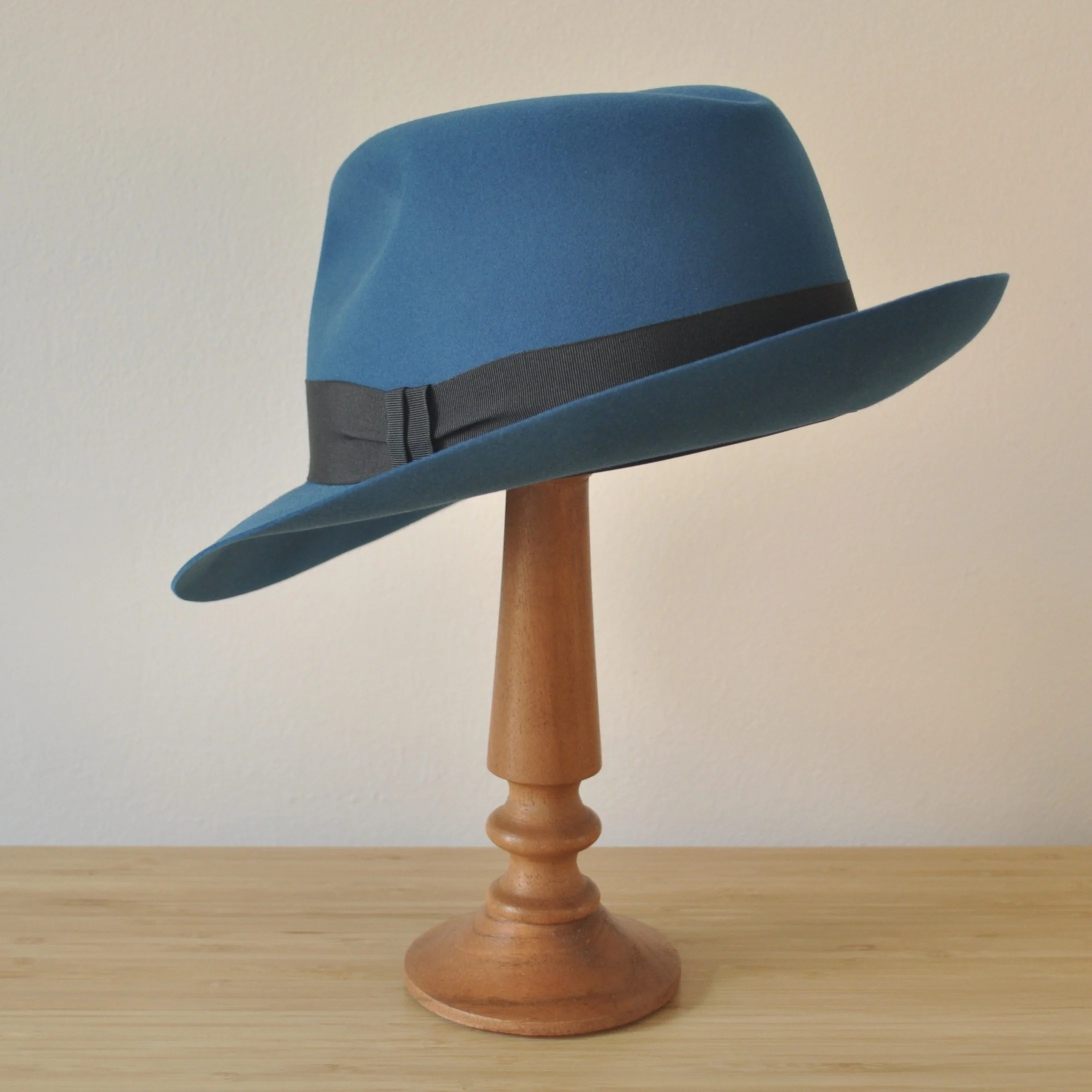 Three-quarter back left view of a cobalt blue felt fedora with a softly pinched centre dent crown, medium snap brim, and very dark blueish grey grosgrain ribbon with a modern tie (no bow).