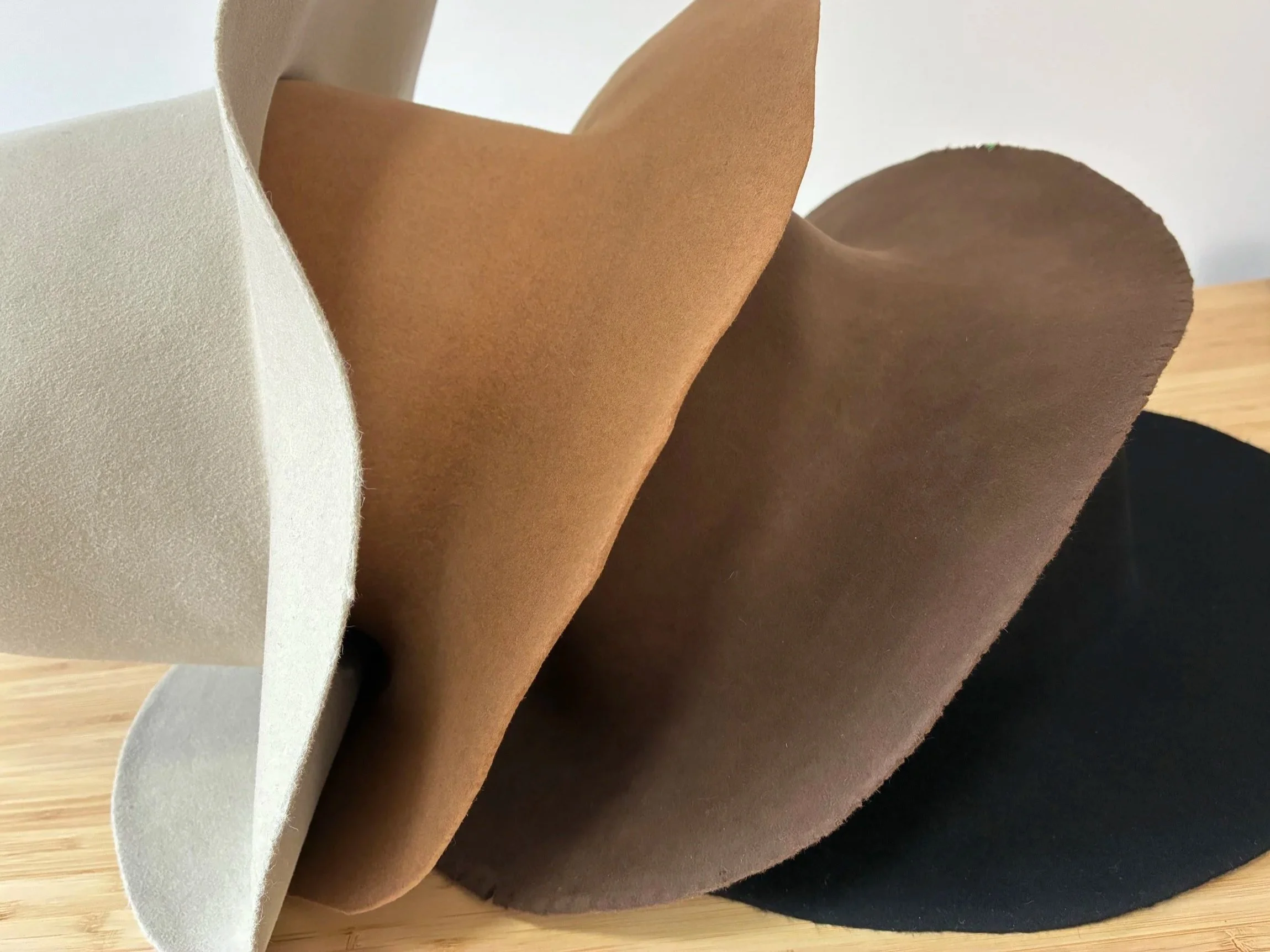 Stacked felt hat bodies in cream, brown, and black colors on a wooden surface.
