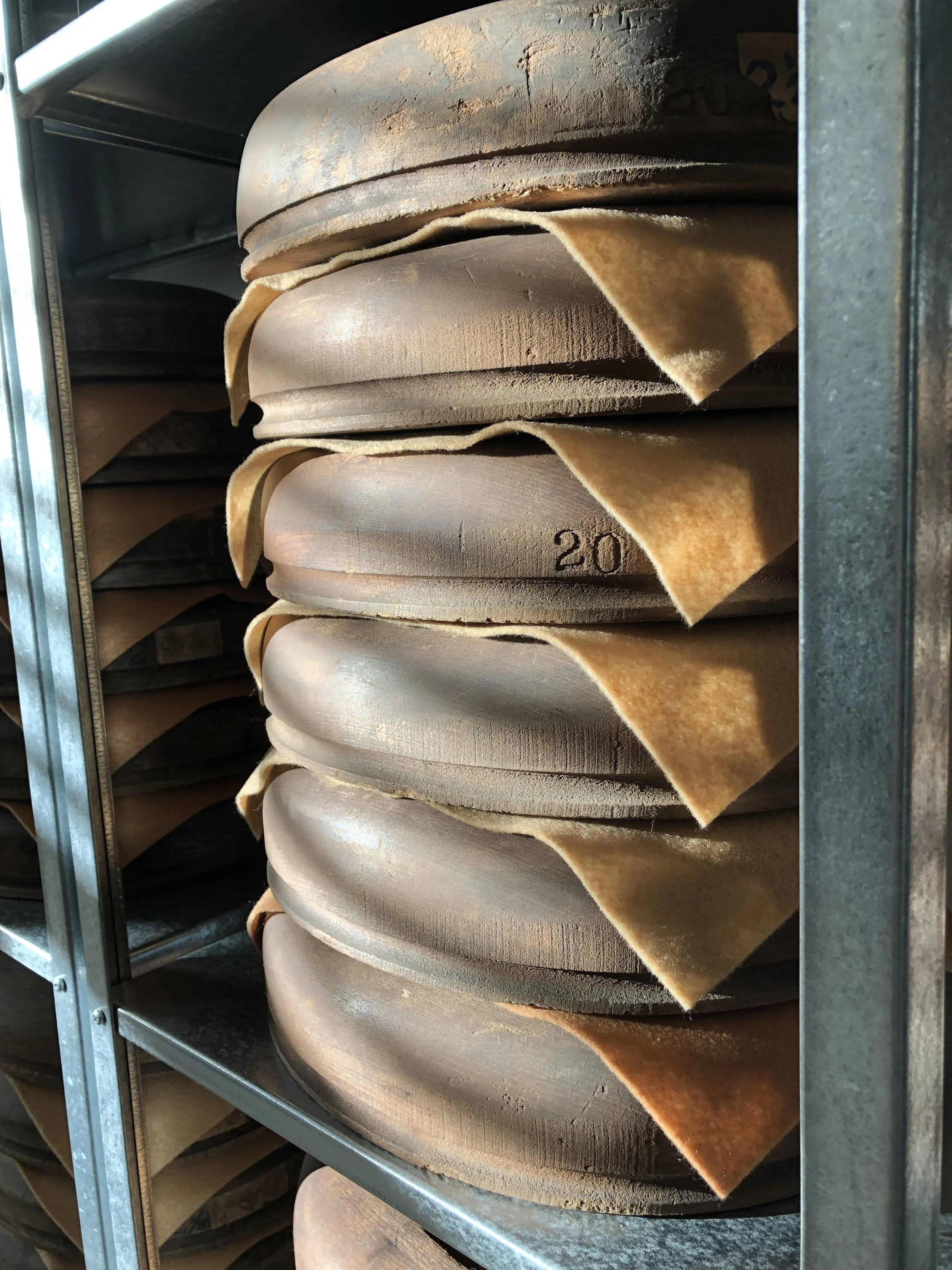 Stacks of wooden hat brim flanges stored on metal shelves, with felt sheets as separators, illuminated by diffuse light passing through blinds.
