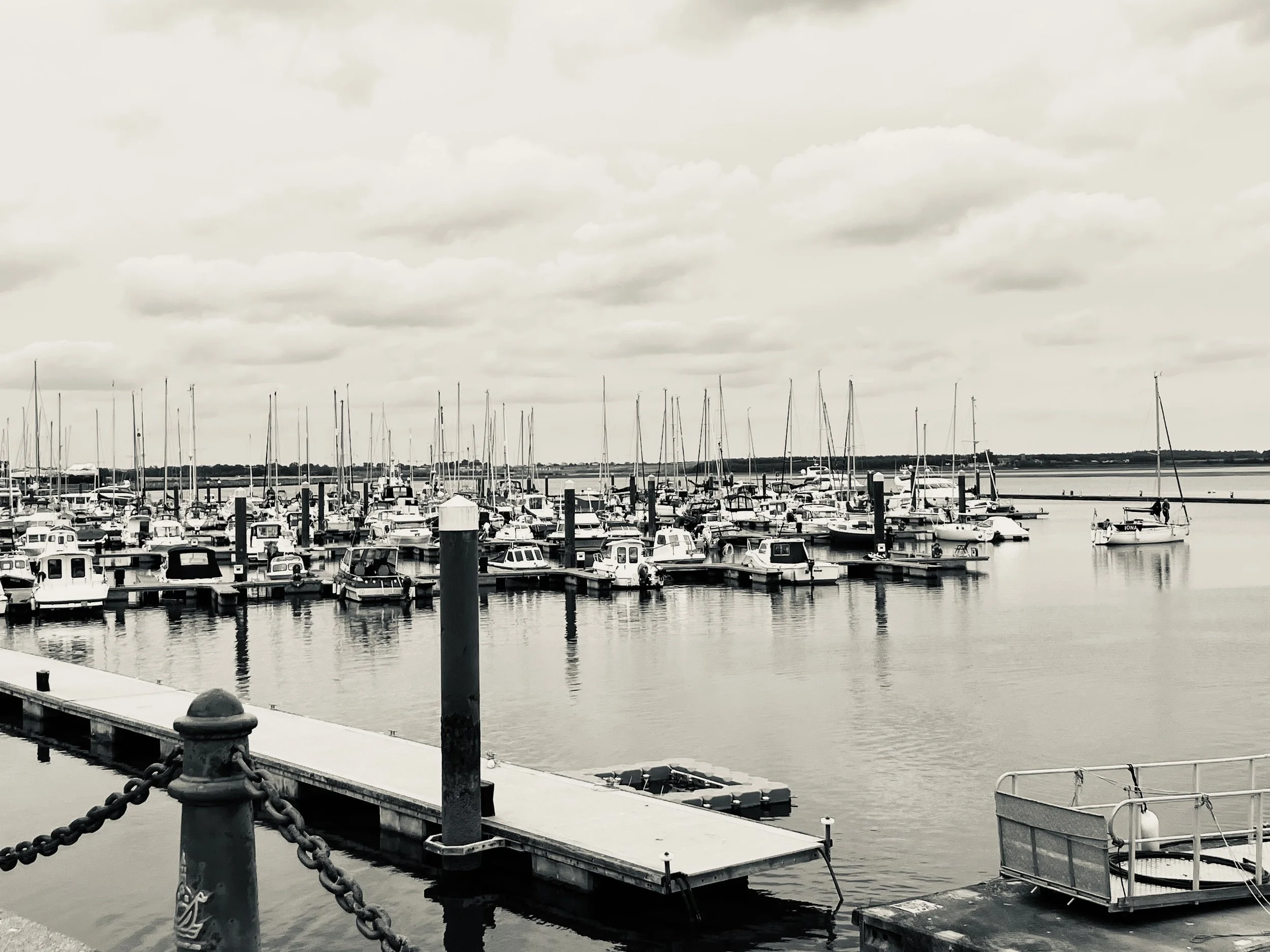 Black and white photo of a marina with numerous boats and yachts docked on calm water under a cloudy sky.