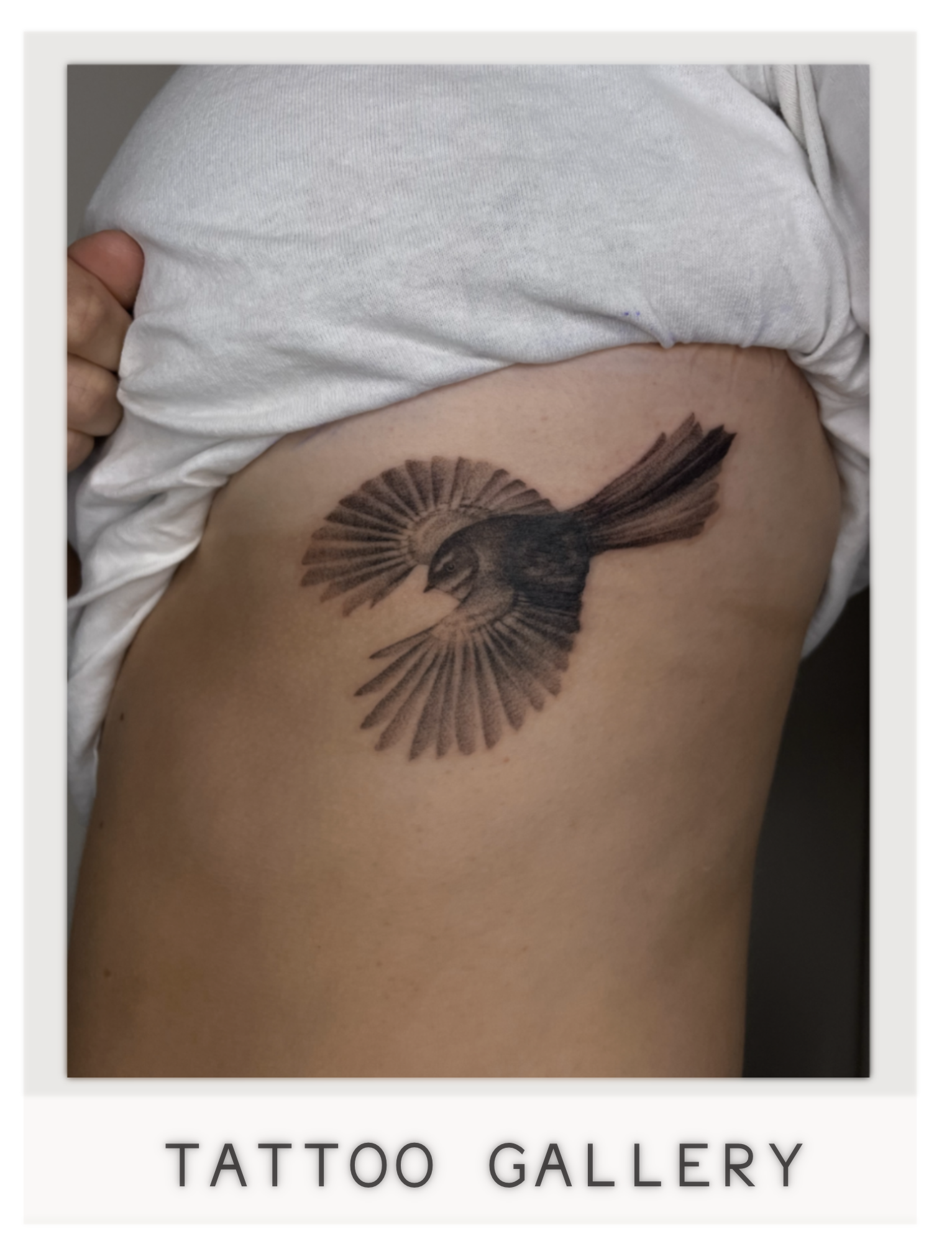 Tattoo of a bird with outstretched wings on person's side, partially covered by a white shirt.