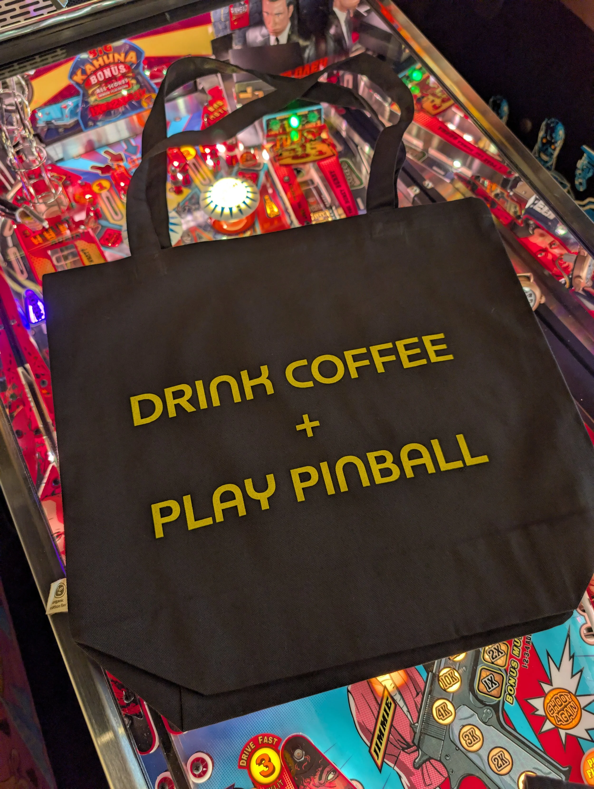 Black tote bags laying on pinball machine highlighting side showing slogan of "Drink Coffee + Play Pinball"