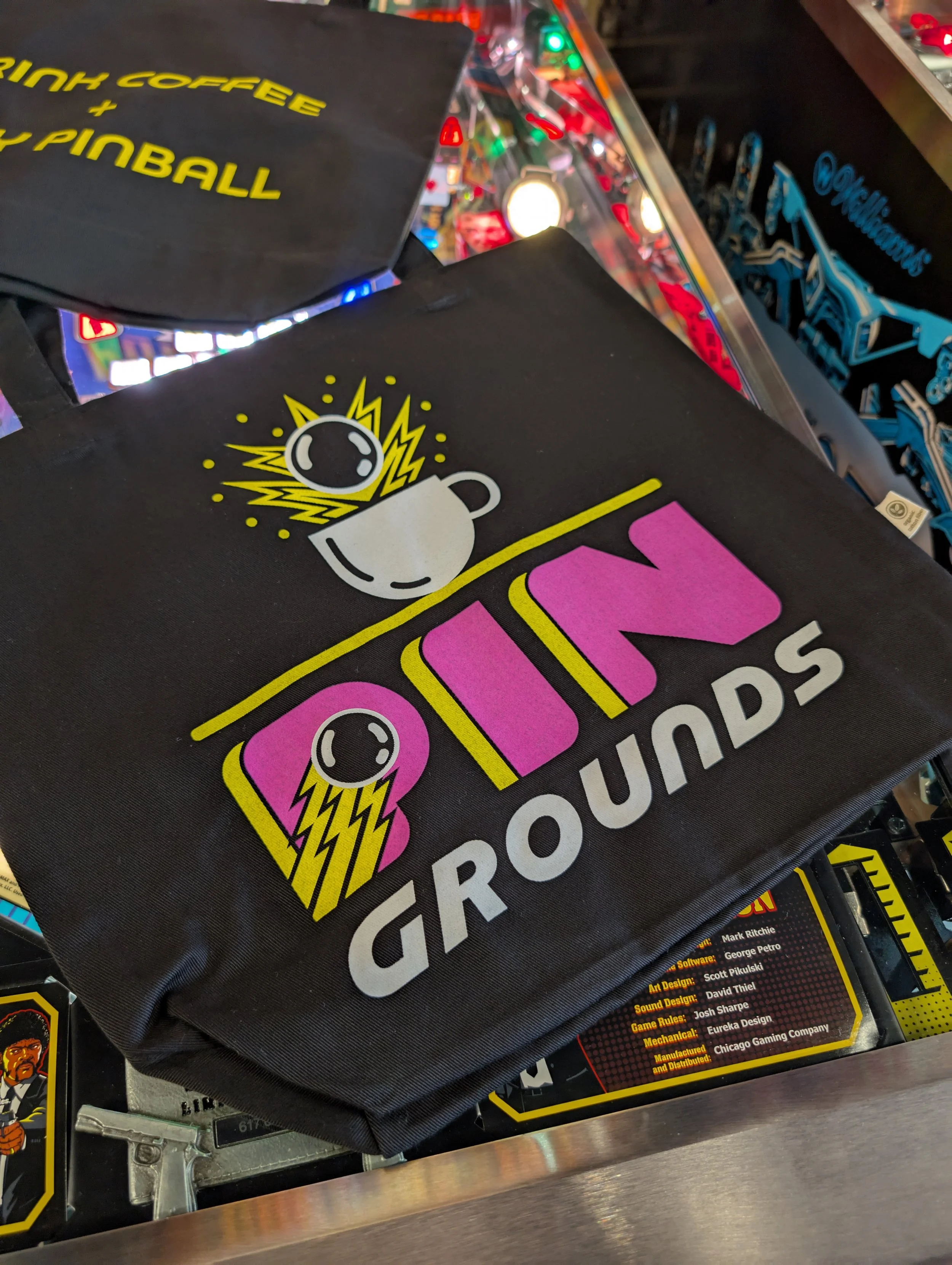 Black tote bags laying on pinball machine highlighting side showing Pin Grounds Coffee logo