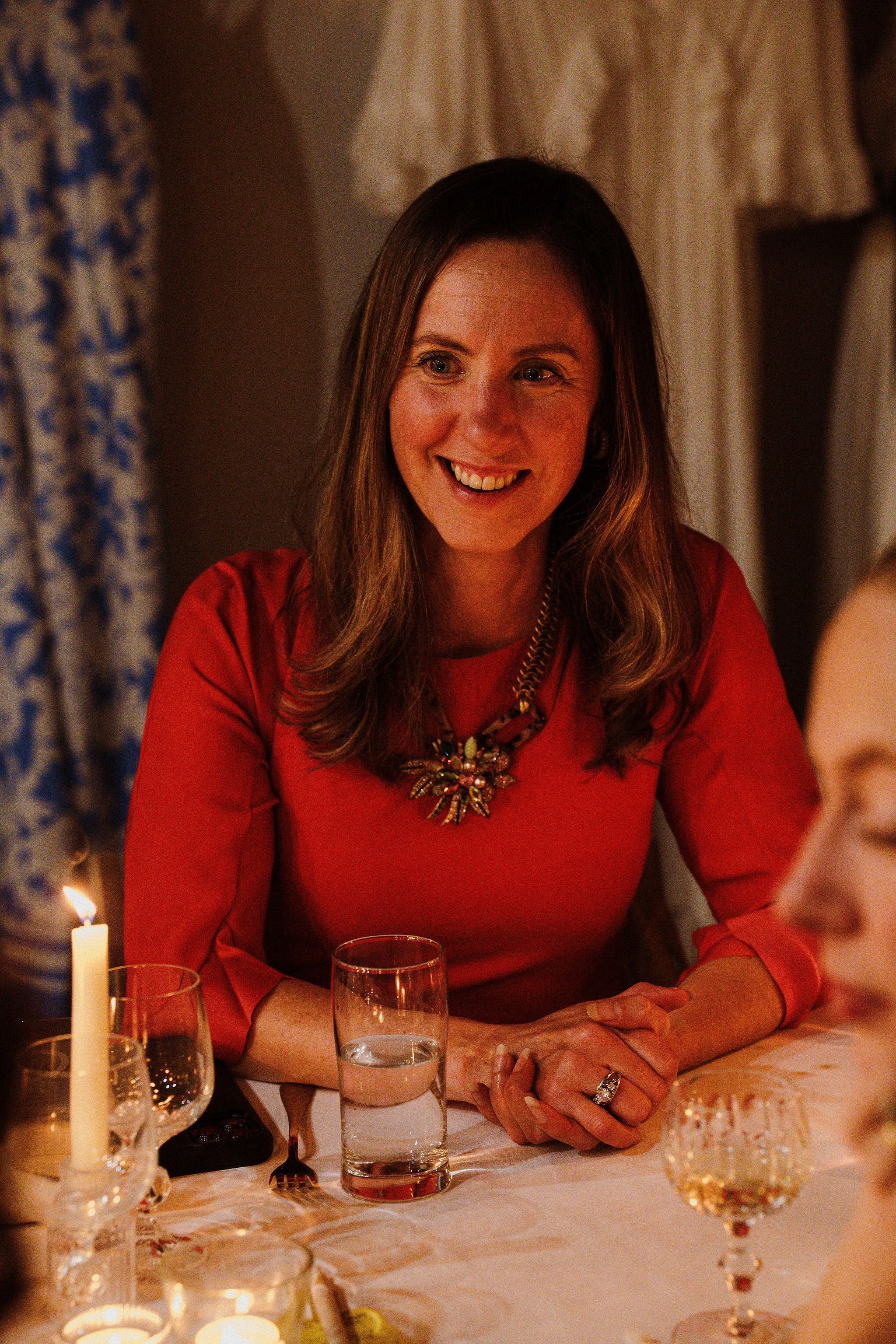 A woman with long brown hair smiling while sitting at a dinner table in a warmly lit room, wearing a red dress and a decorative necklace.