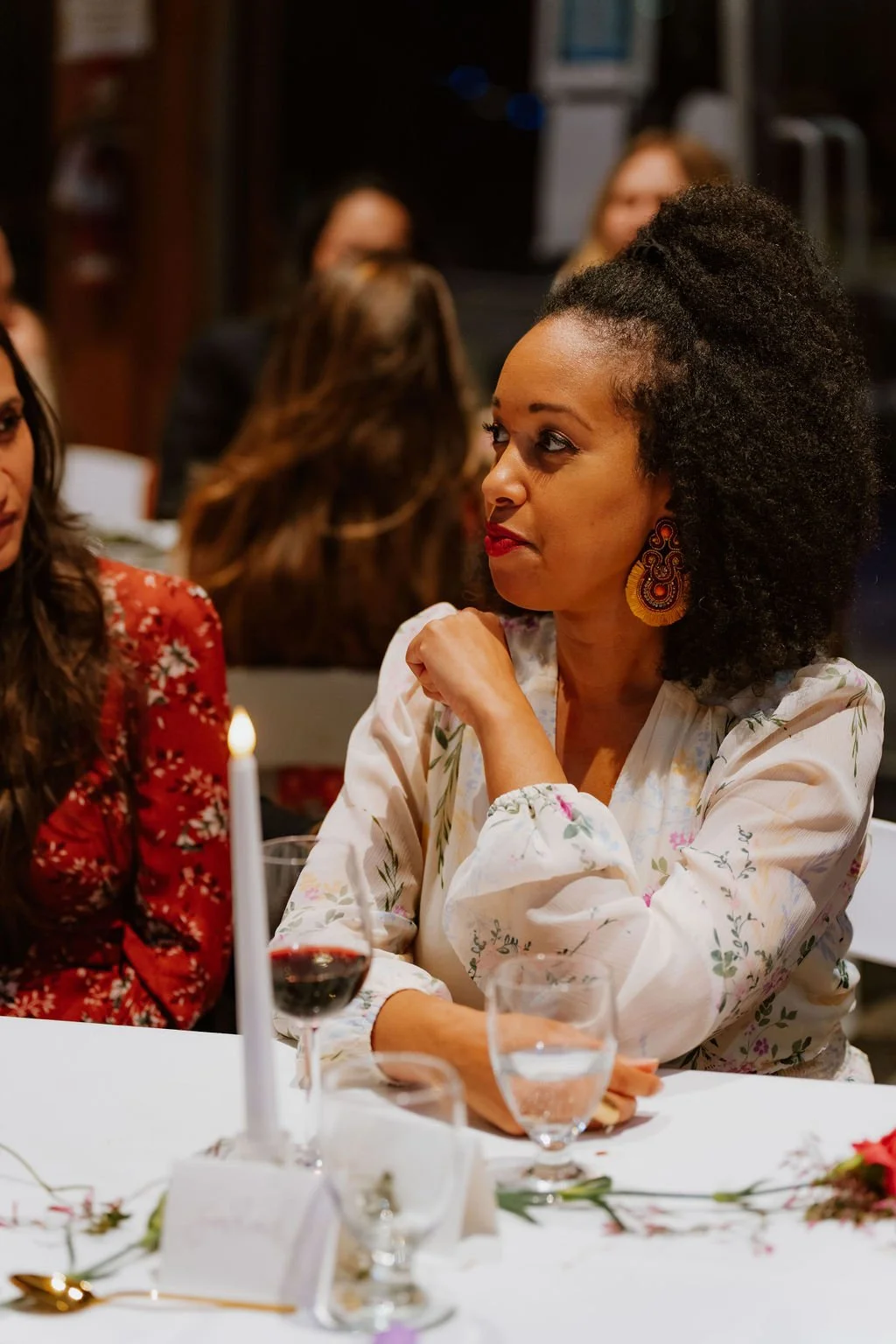 A woman with curly black hair and red lipstick sitting at a table with wine and water glasses, wearing a white floral blouse and large colorful earrings, engaged in conversation at a social gathering.