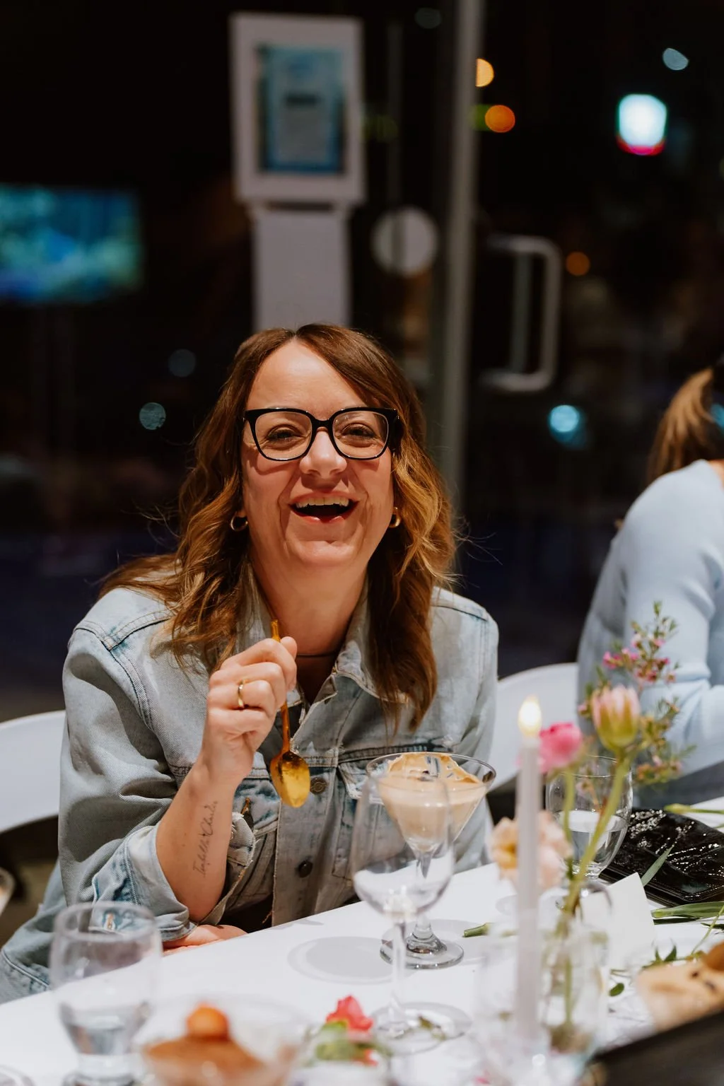 A woman with shoulder-length brown hair and glasses, smiling and holding a spoon, sitting at a decorated table with desserts and floral arrangements in a festive setting.