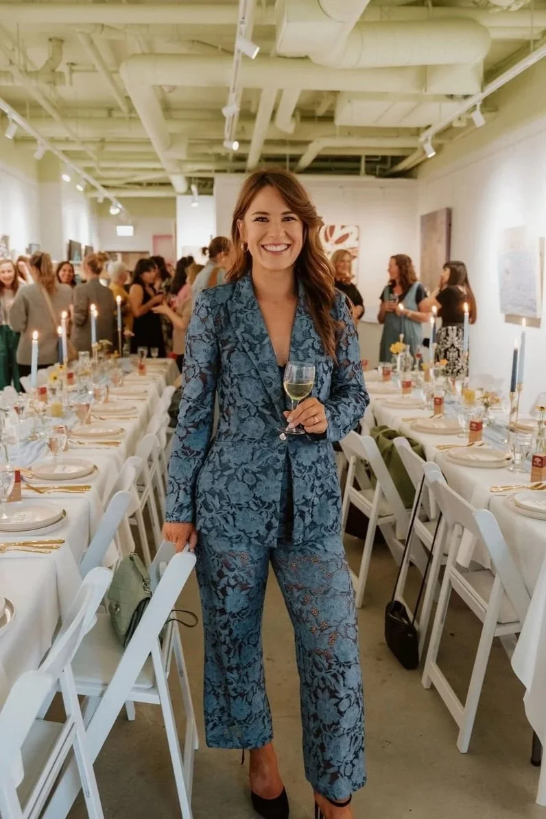 A woman in a blue lace suit smiling and holding a glass of white wine at a banquet with decorated tables and guests in the background.