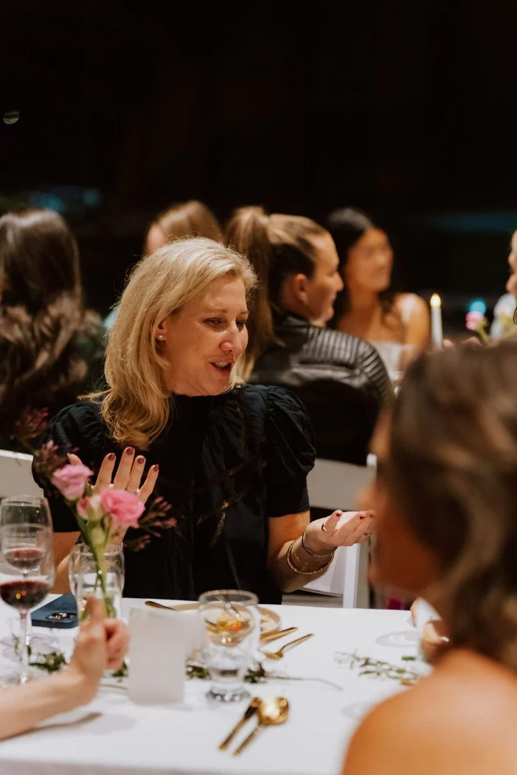 A group of women sitting at a dinner table during a social event, with one woman in the center smiling and talking, surrounded by wine glasses, floral arrangements, and tableware.