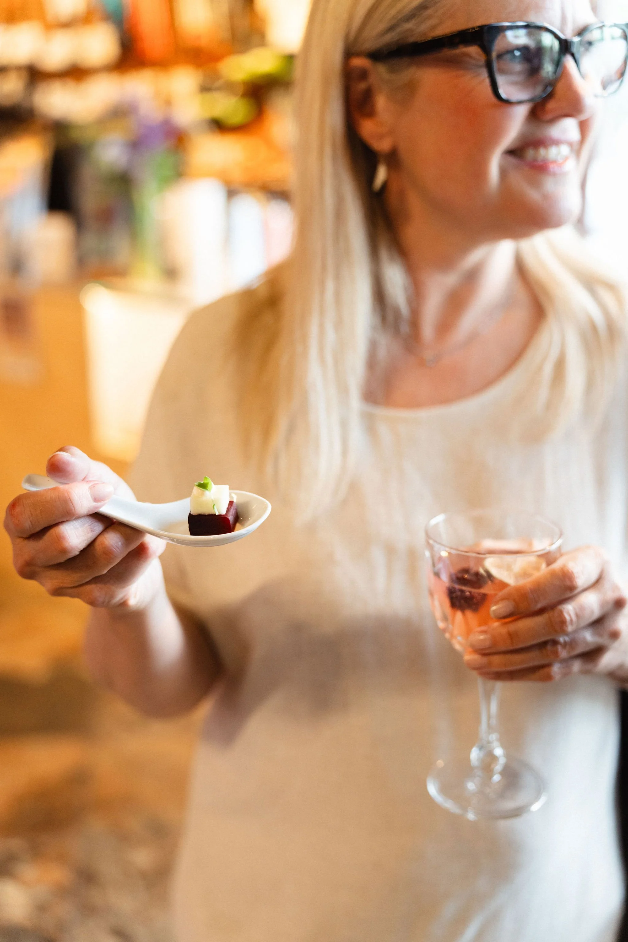 A woman with blonde hair and glasses holding a glass of pink drink and a spoon with a small piece of dessert in a cozy indoor setting.