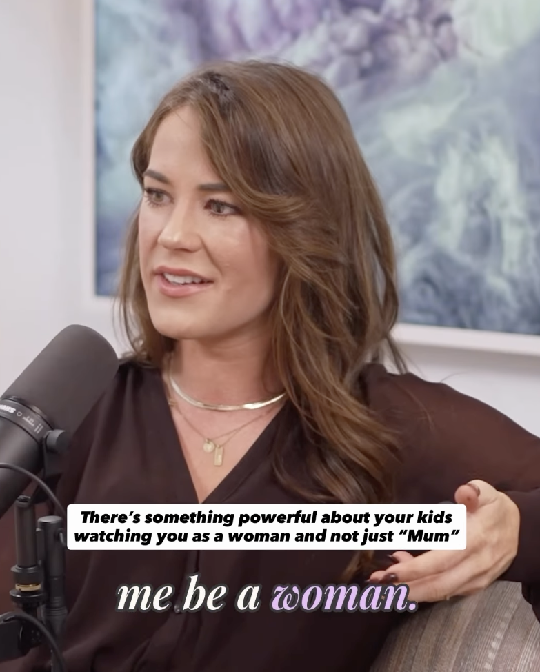 A woman with wavy brown hair and light makeup, wearing a black top and layered necklaces, is speaking into a microphone against a colorful abstract background. Subtitles read: "There’s something powerful about your kids watching you as a woman and not just ‘Mum’" and "me be a woman."