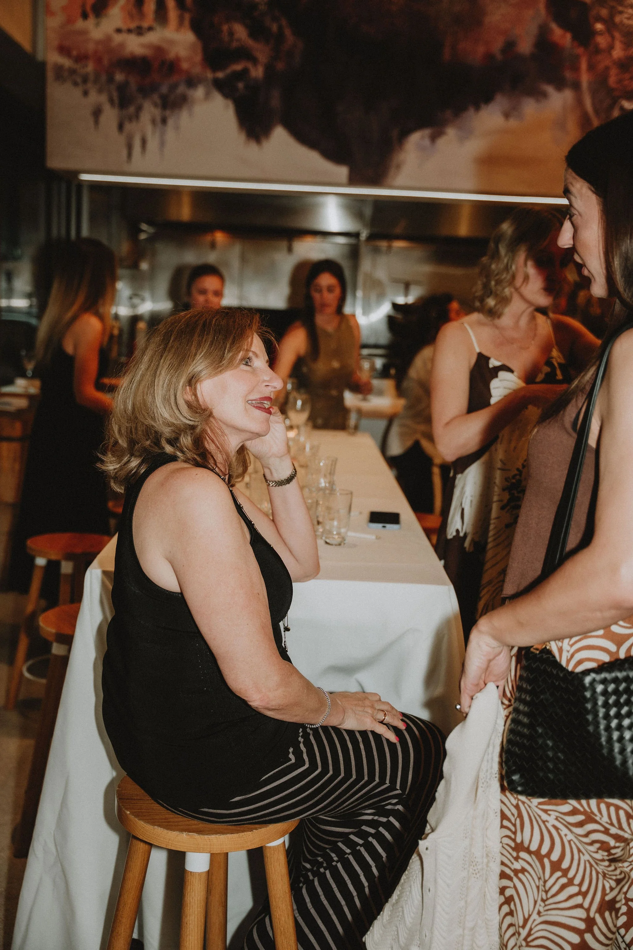 A woman with blonde hair wearing a black sleeveless top and striped pants sitting on a wooden stool in a restaurant or bar, engaged in conversation with another woman standing nearby.