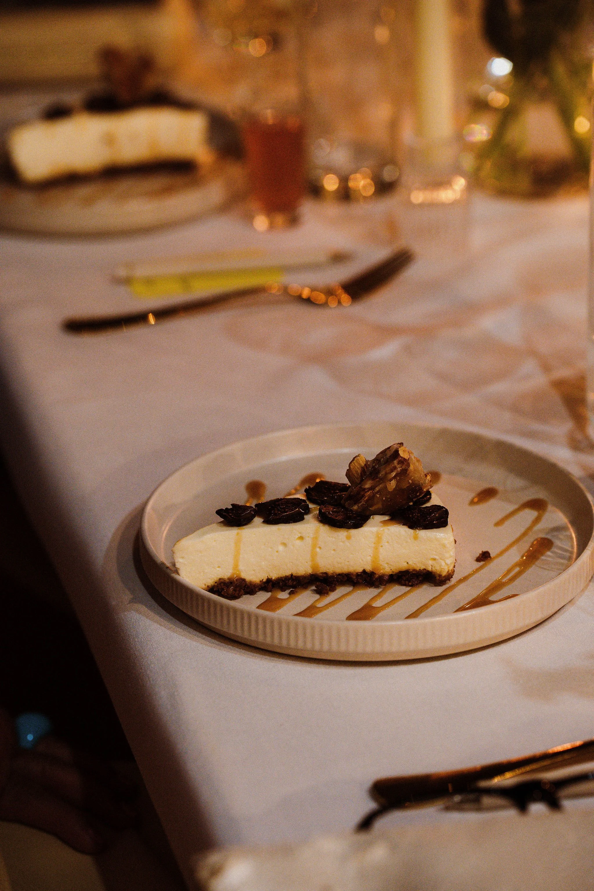 A slice of cheesecake topped with a walnut and raisins, with caramel drizzle on a white plate at an elegantly set dinner table.