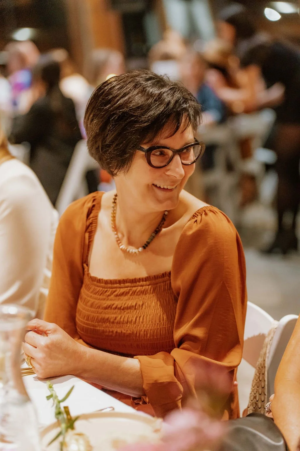 A woman with short dark hair, glasses, and a warm brown dress sitting at a festive dinner table, smiling and engaging in conversation, with a lively gathering of people in the background.