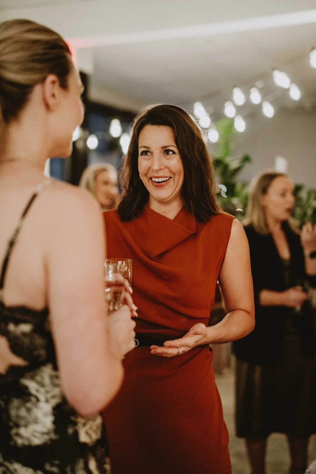 A woman with shoulder-length dark hair wearing a sleeveless red dress is smiling and talking to another woman holding a glass at a social event. Other women are in the background in an indoor setting with string lights.