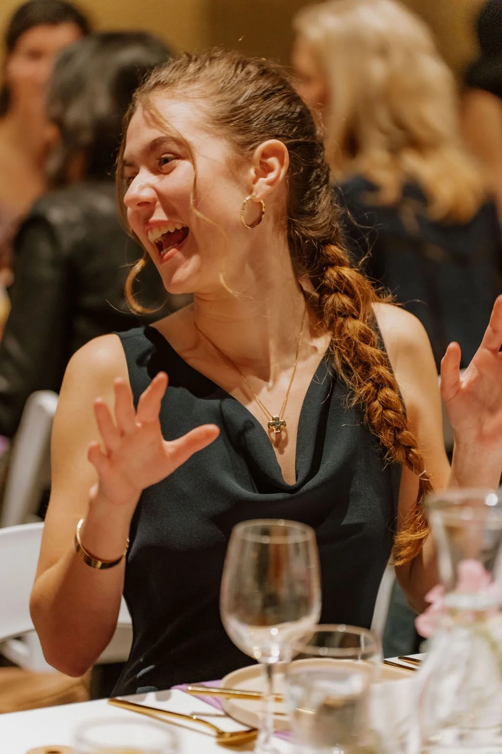 A woman laughing and gesturing with her hands during a social gathering, seated at a table with glassware and table settings.