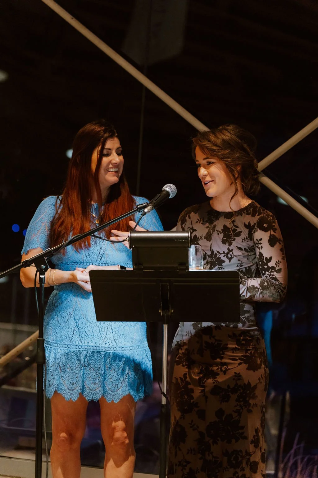 Two women with microphones standing at a podium, smiling and speaking at an event in a dimly lit indoor setting.