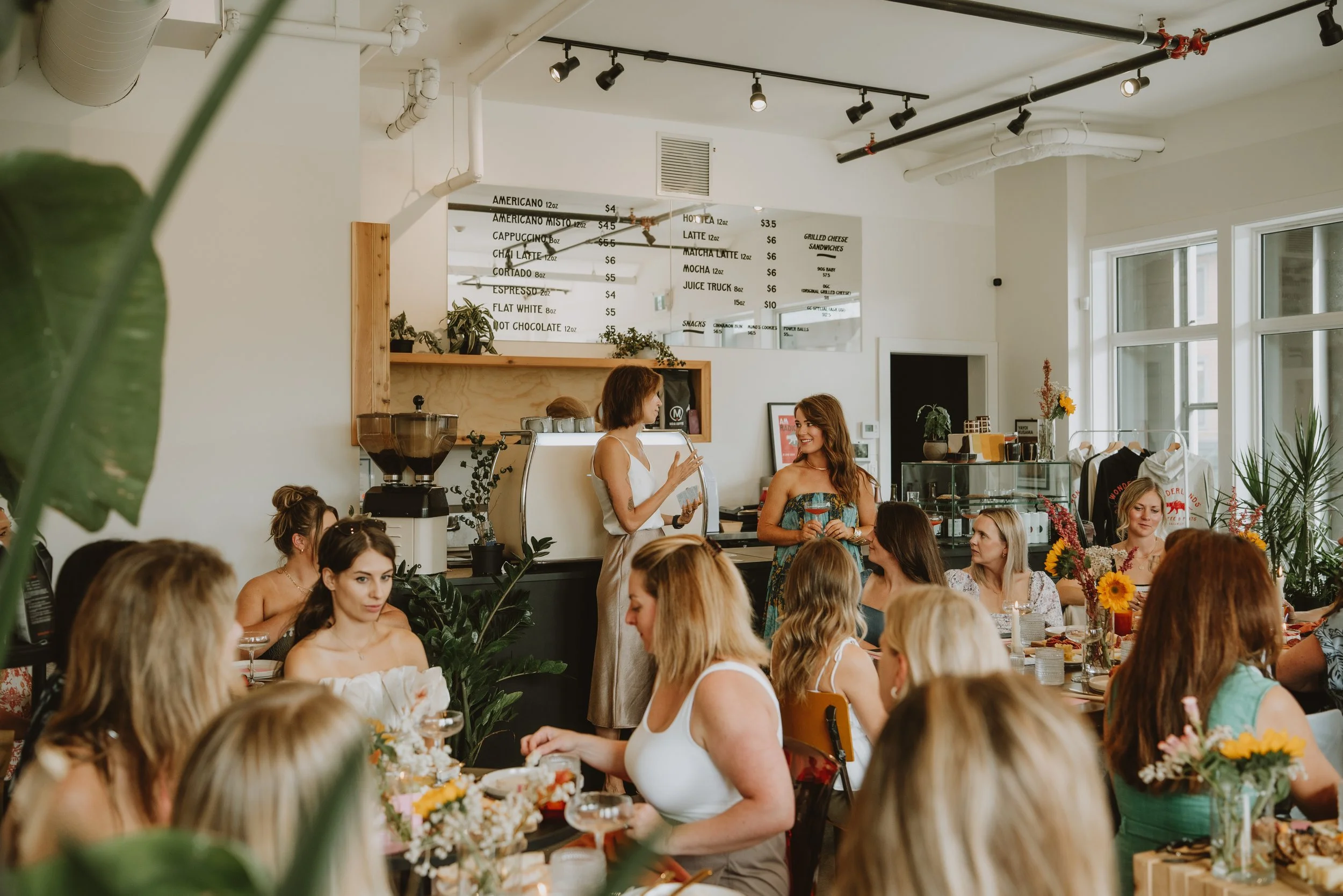 Women gathered in a cafe for a meeting or celebration, with two women standing and smiling at each other in front of a group of seated women at tables decorated with flowers, inside a bright, modern cafe with a menu above the counter.
