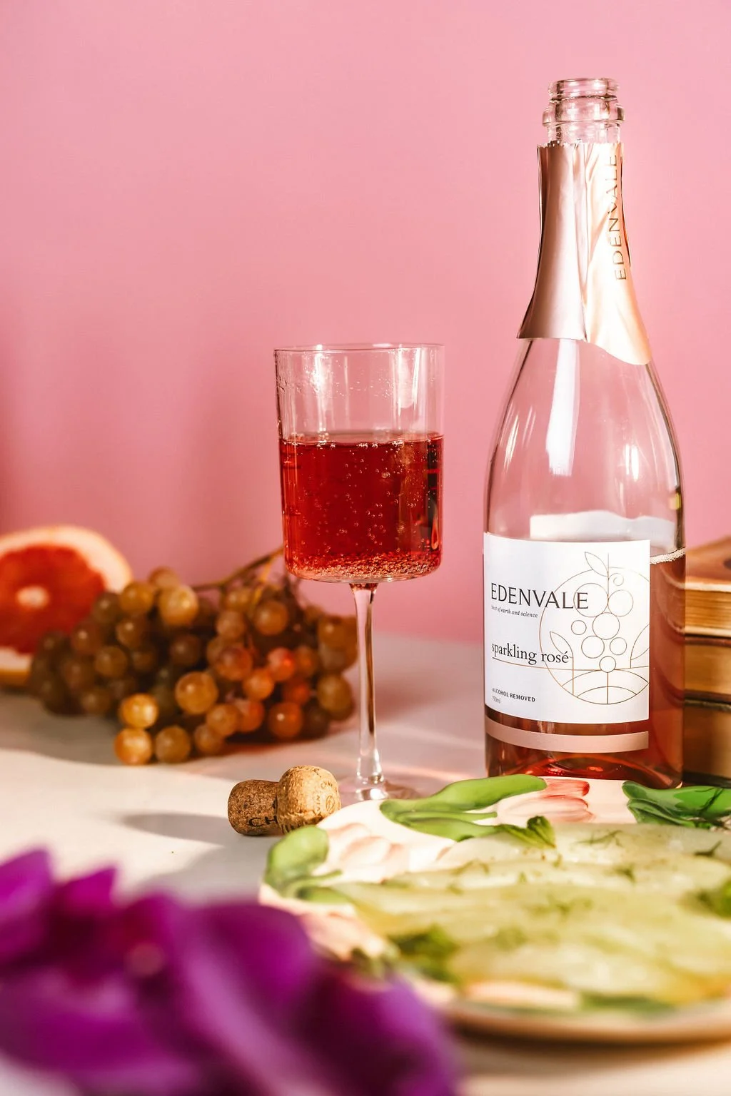 A glass of sparkling rosé wine, a bottle of Edenvale sparkling rosé, grapes, a grapefruit, a cork, a plate of cucumber slices with herbs, and purple flowers on a table with a pink background.