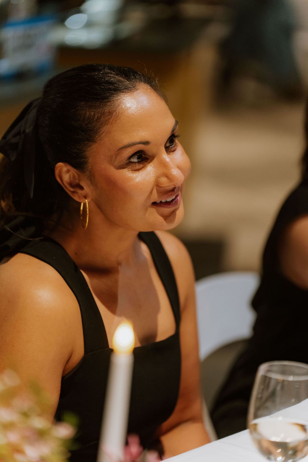 A woman with dark hair tied back, wearing gold hoop earrings and a black sleeveless top, smiling and looking to her left at a dinner table.