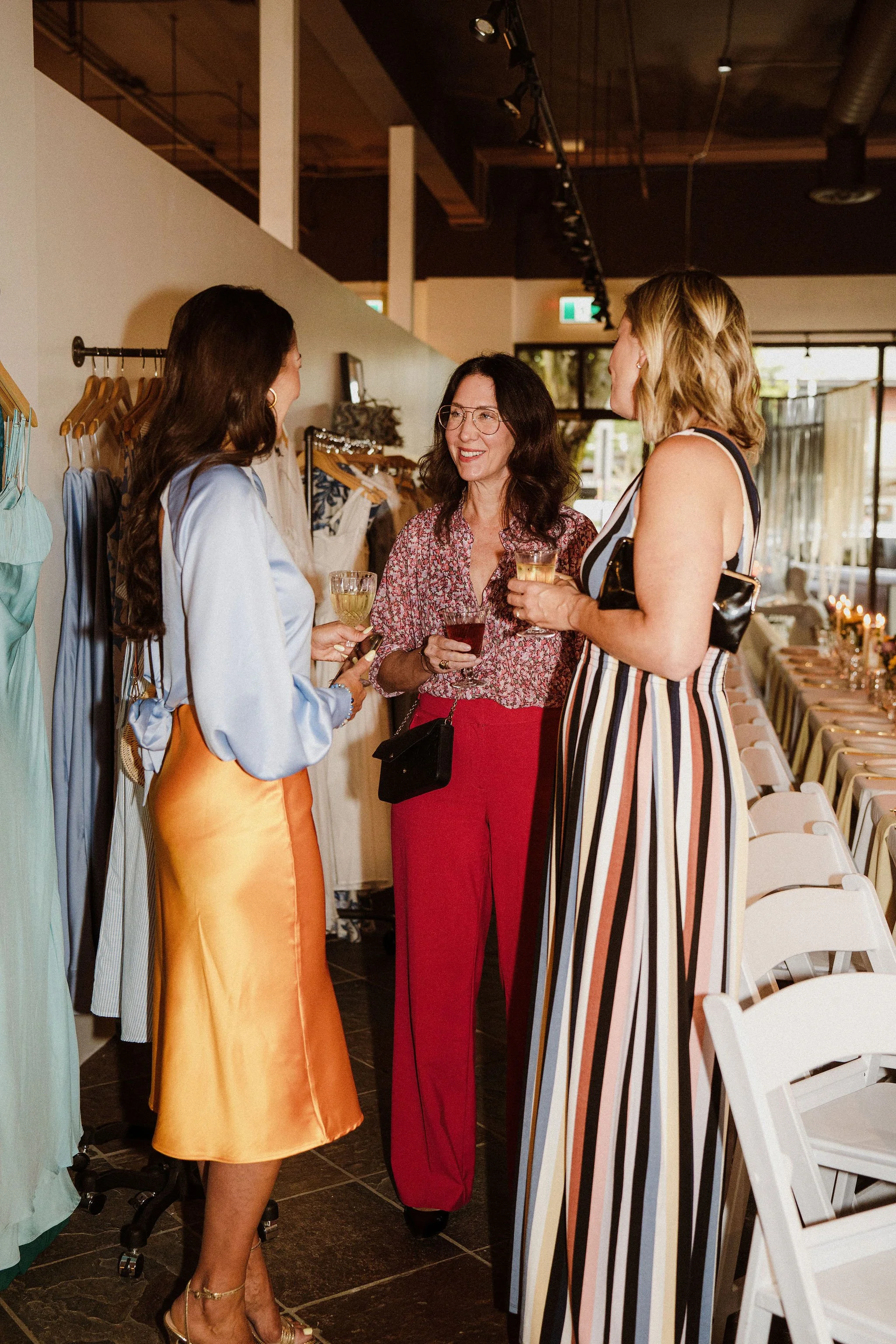 Three women chatting and holding drinks in a store or restaurant with a long table set for a meal in the background.